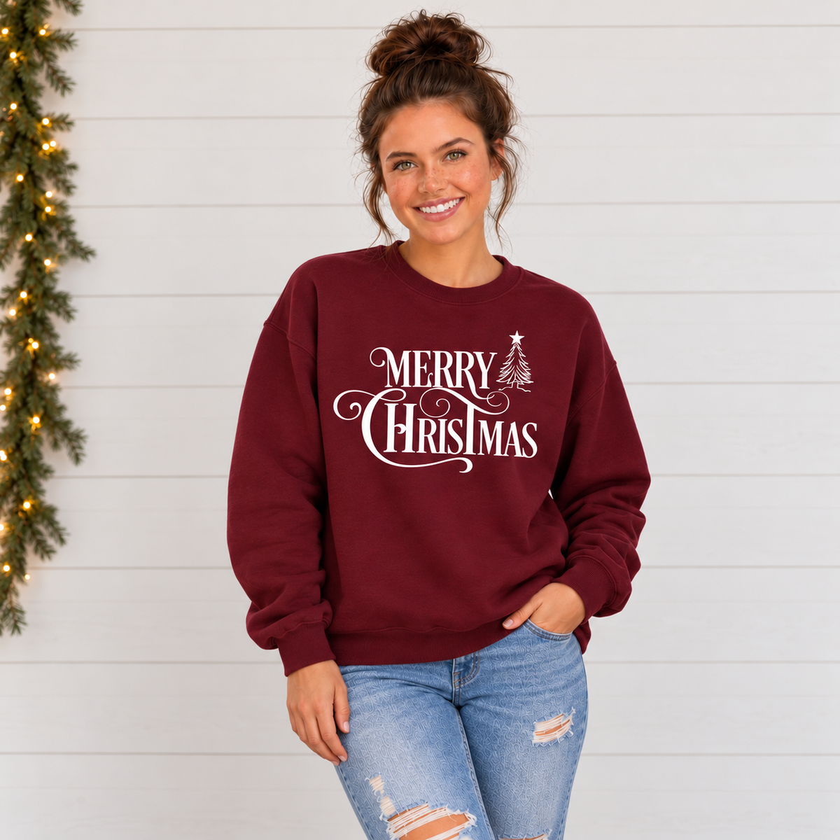 Smiling model in a maroon Merry Christmas crewneck sweatshirt against a clean white shiplap wall, showing the white script and star-topped tree.