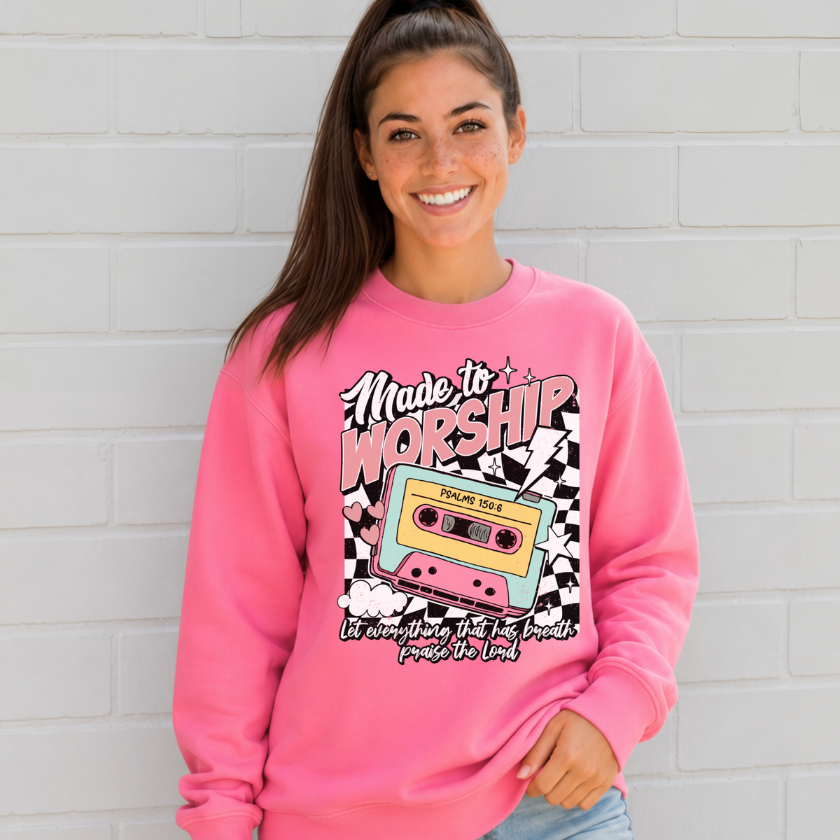 Woman wearing a Bright Pink Made to Worship crewneck sweatshirt with a retro cassette graphic against a white brick wall.