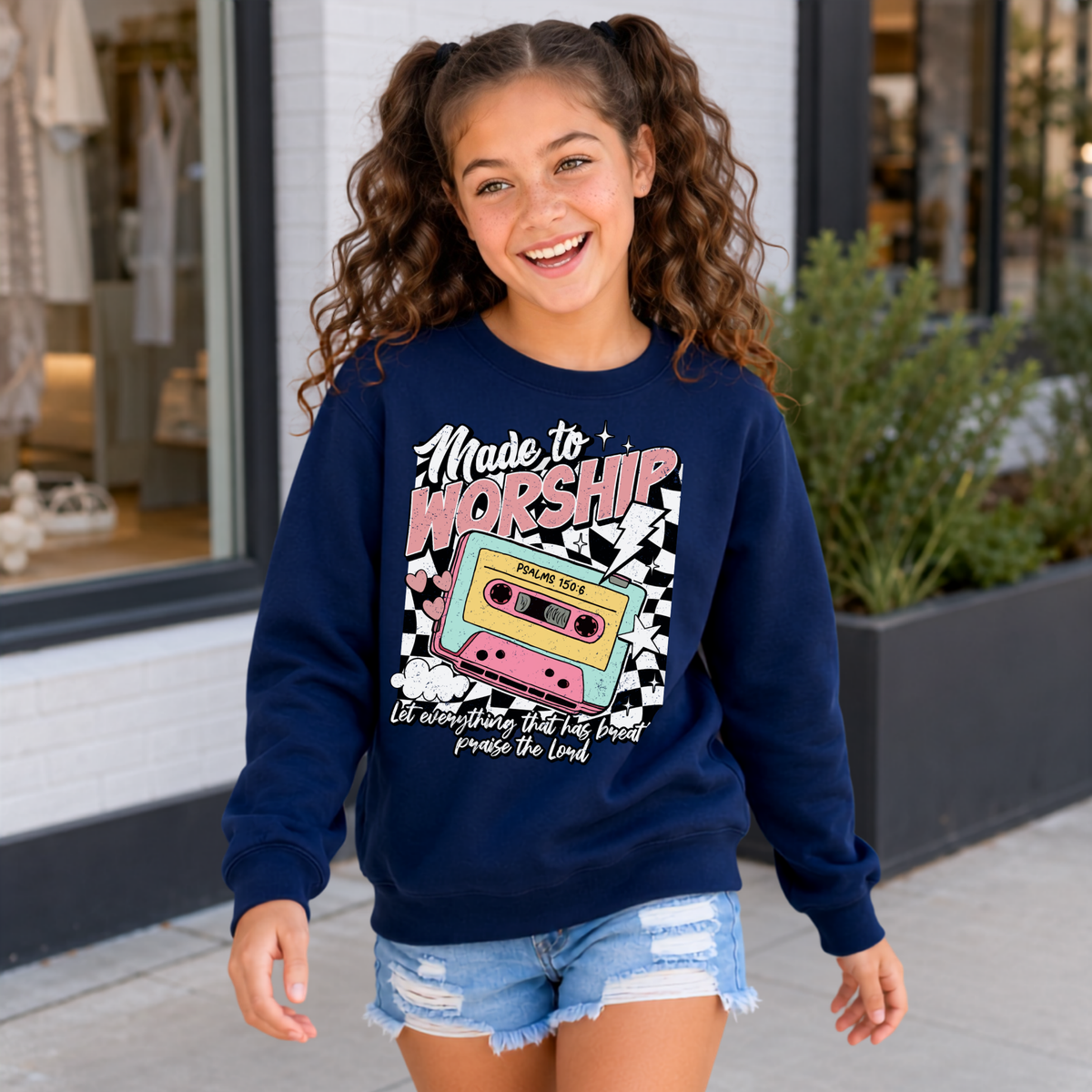 Youth girl wearing a navy Made to Worship crewneck sweatshirt with a colorful retro cassette graphic outside a storefront.