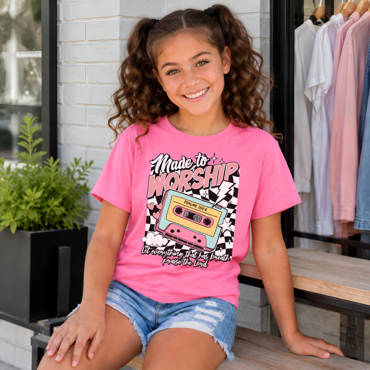 Youth girl wearing a Bright Pink Made to Worship T-shirt with the retro cassette Psalm 150:6 design outside a storefront.
