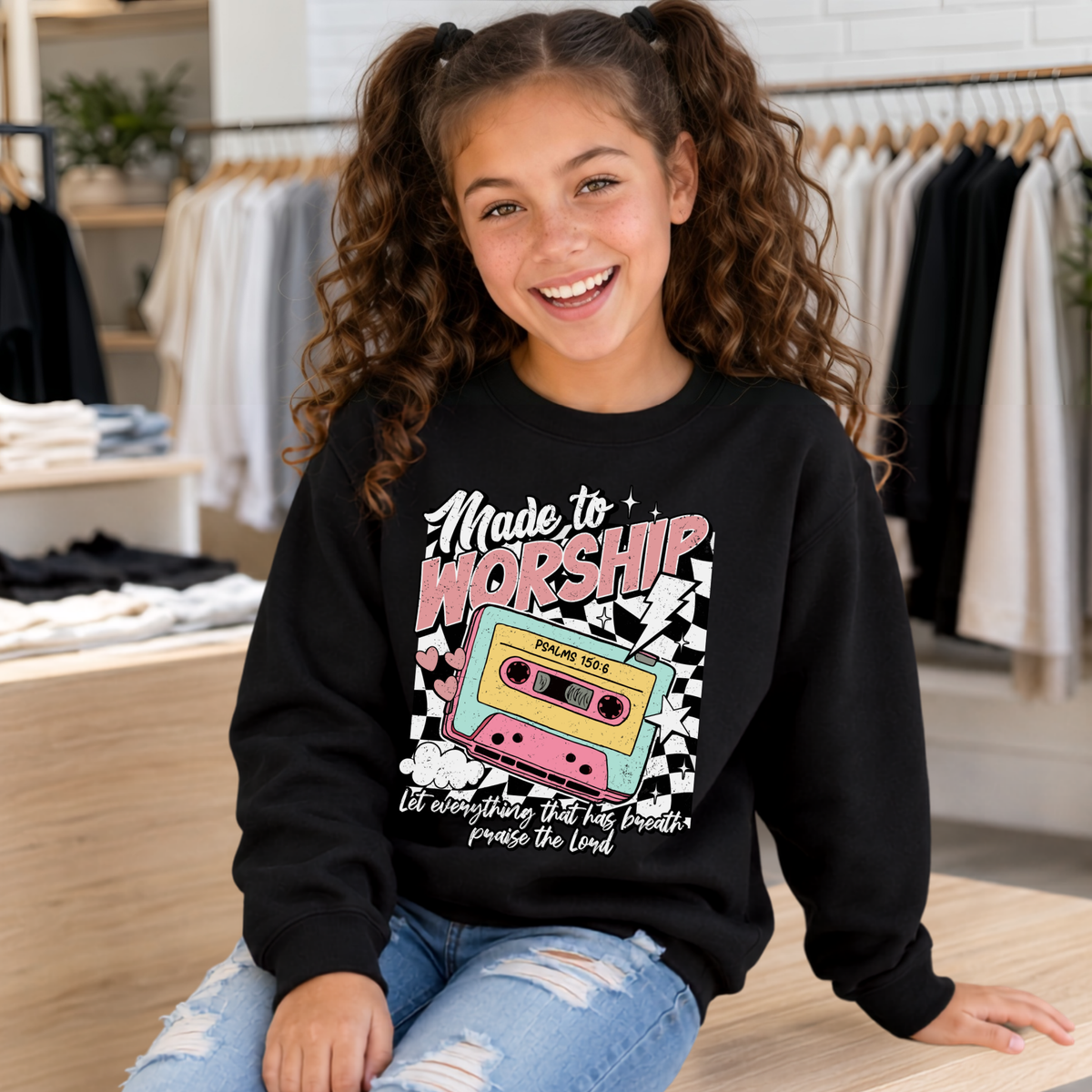 Youth girl wearing a black Made to Worship crewneck sweatshirt with the retro cassette Psalm 150:6 design in a modern clothing store.