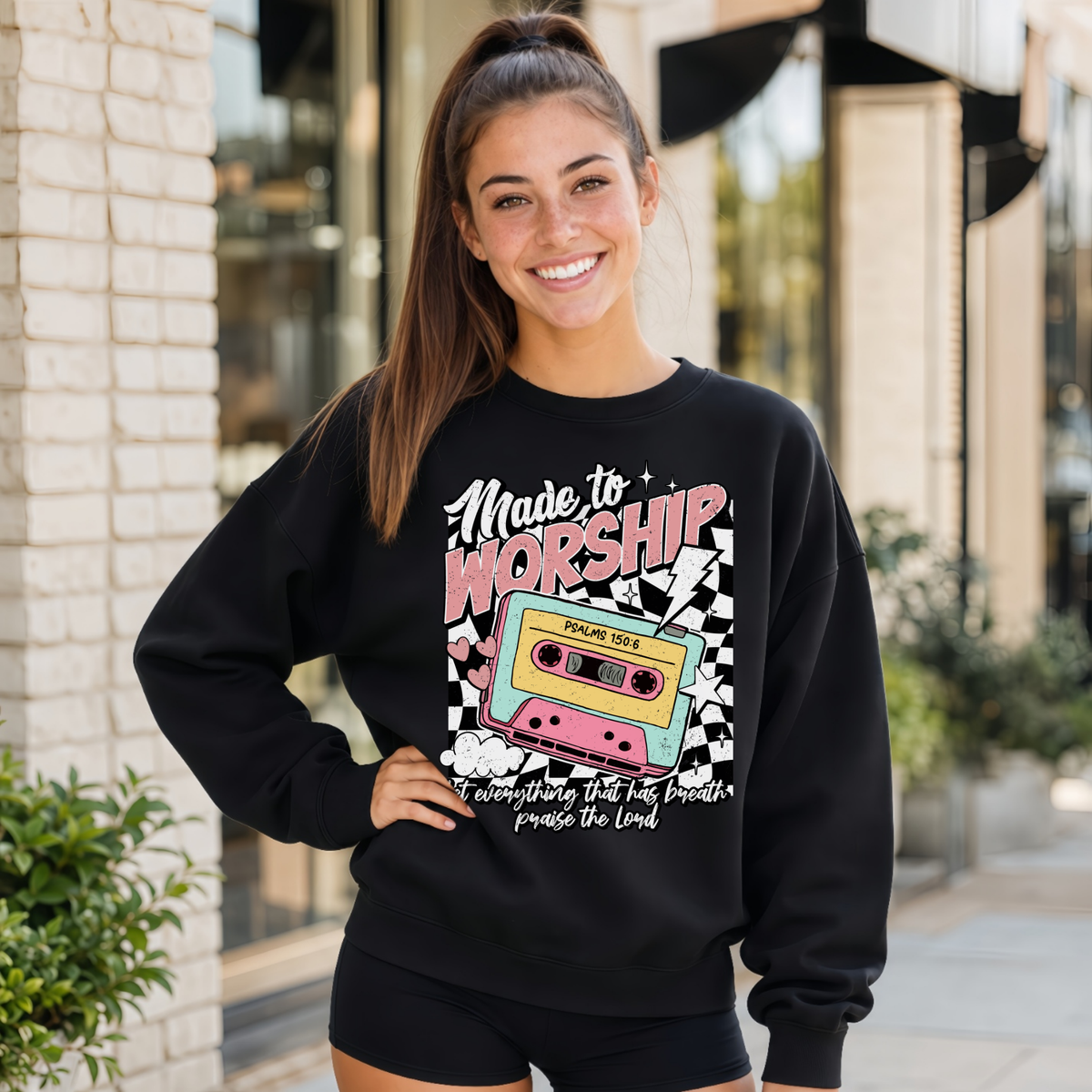Woman wearing a black Made to Worship crewneck sweatshirt with a retro cassette graphic outside a storefront.