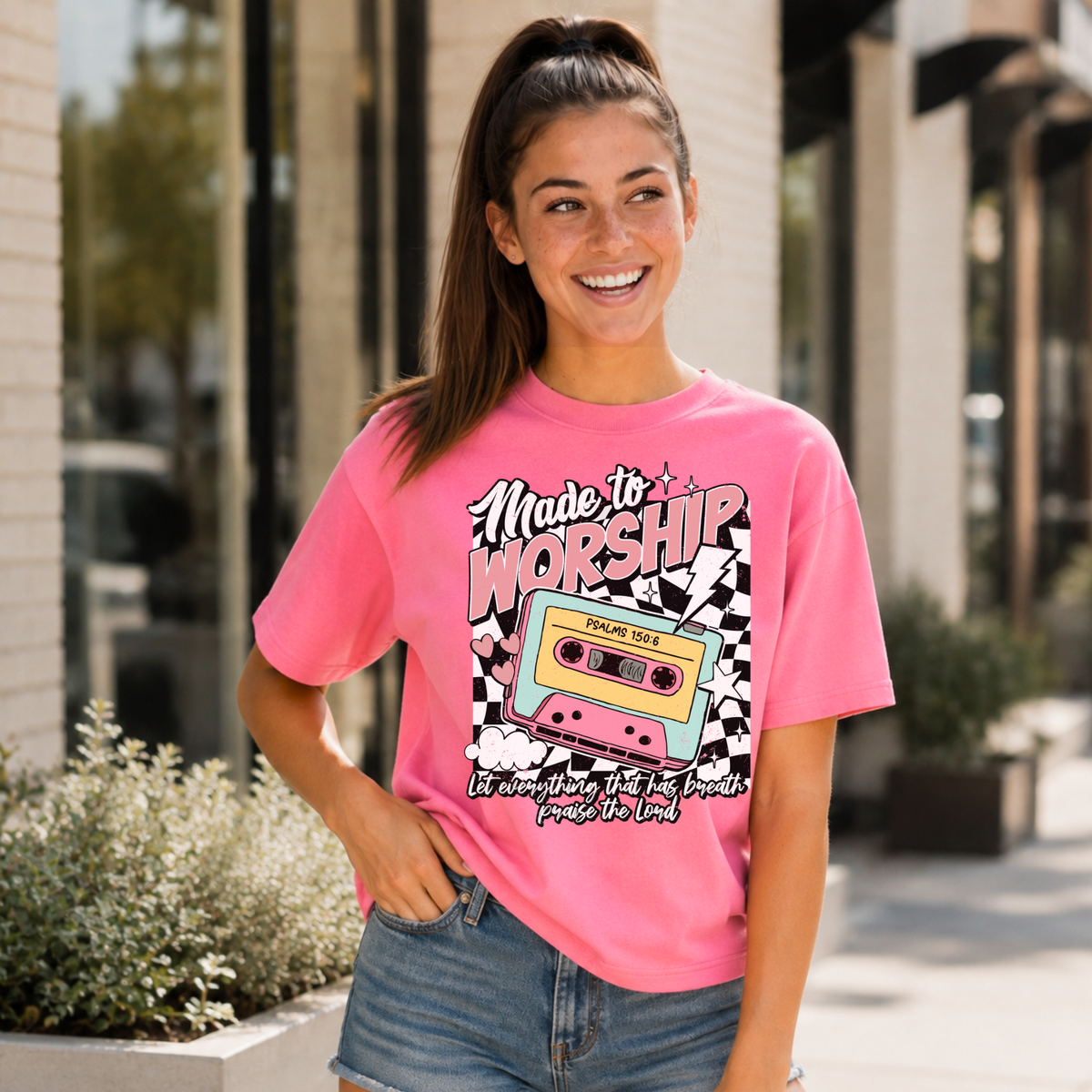 Woman wearing a Bright Pink Made to Worship T-shirt with a retro cassette graphic outside a storefront.