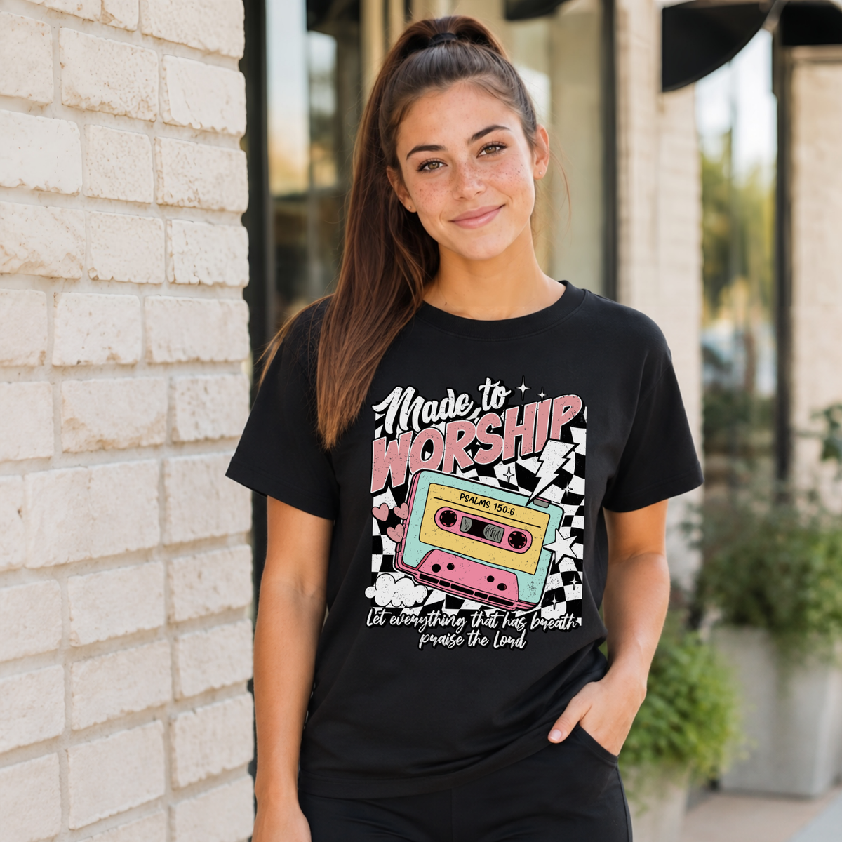 Woman wearing a black Made to Worship T-shirt with a retro cassette graphic outside a storefront.