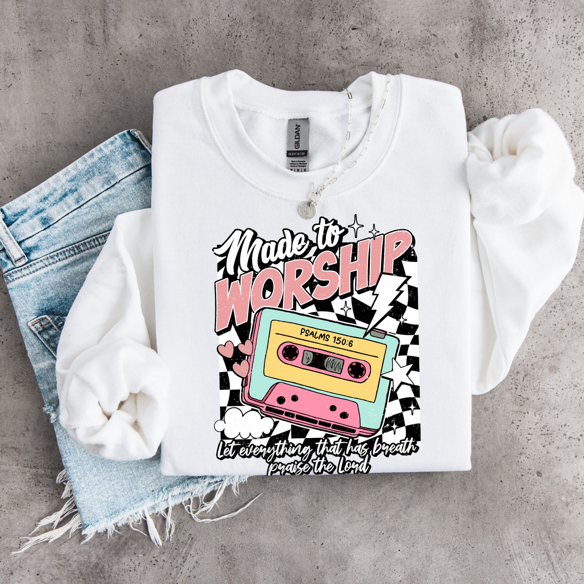 White Made to Worship youth crewneck sweatshirt flat lay with a colorful retro cassette graphic on a gray concrete background.