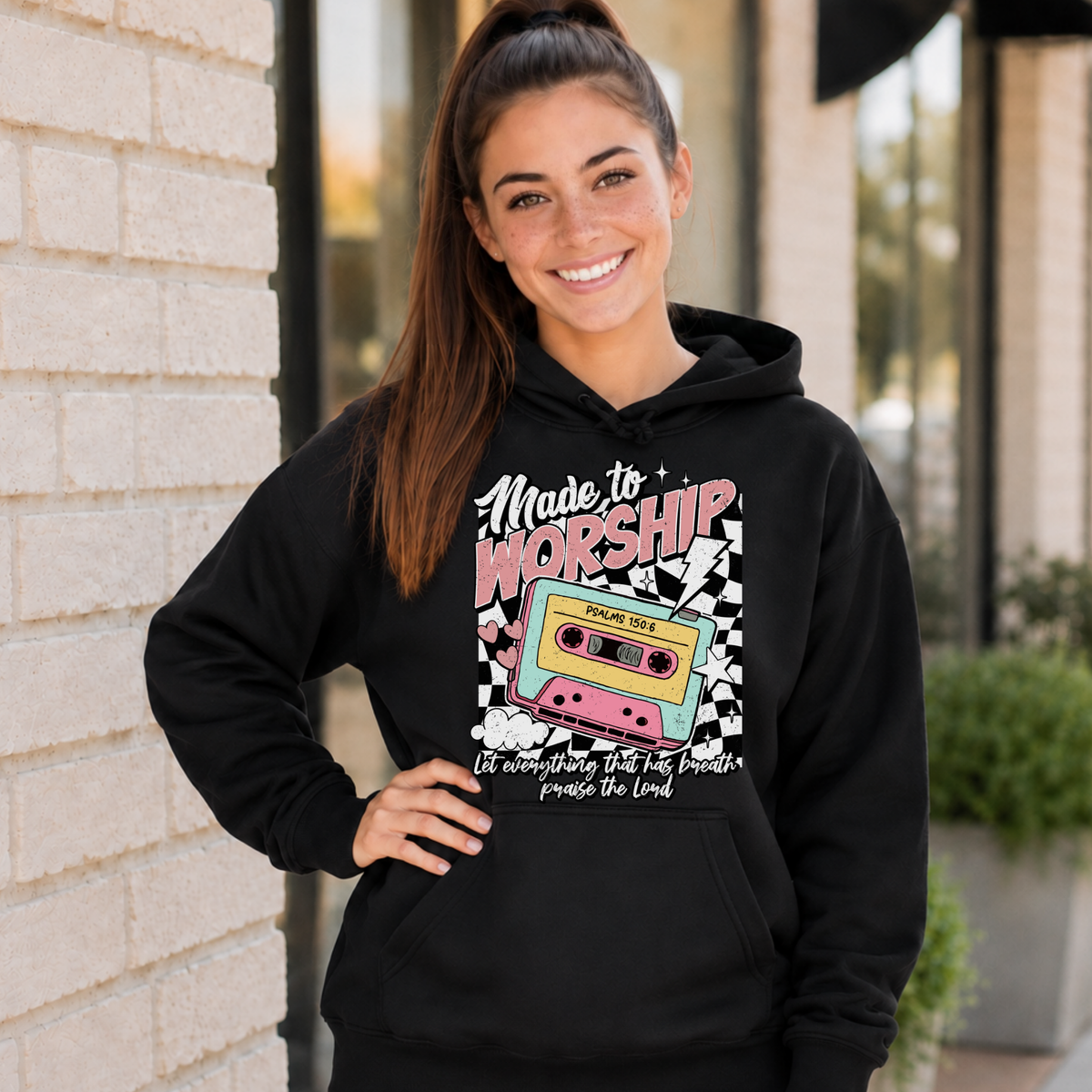 Woman wearing a black Made to Worship hoodie with a retro cassette graphic outside a storefront.