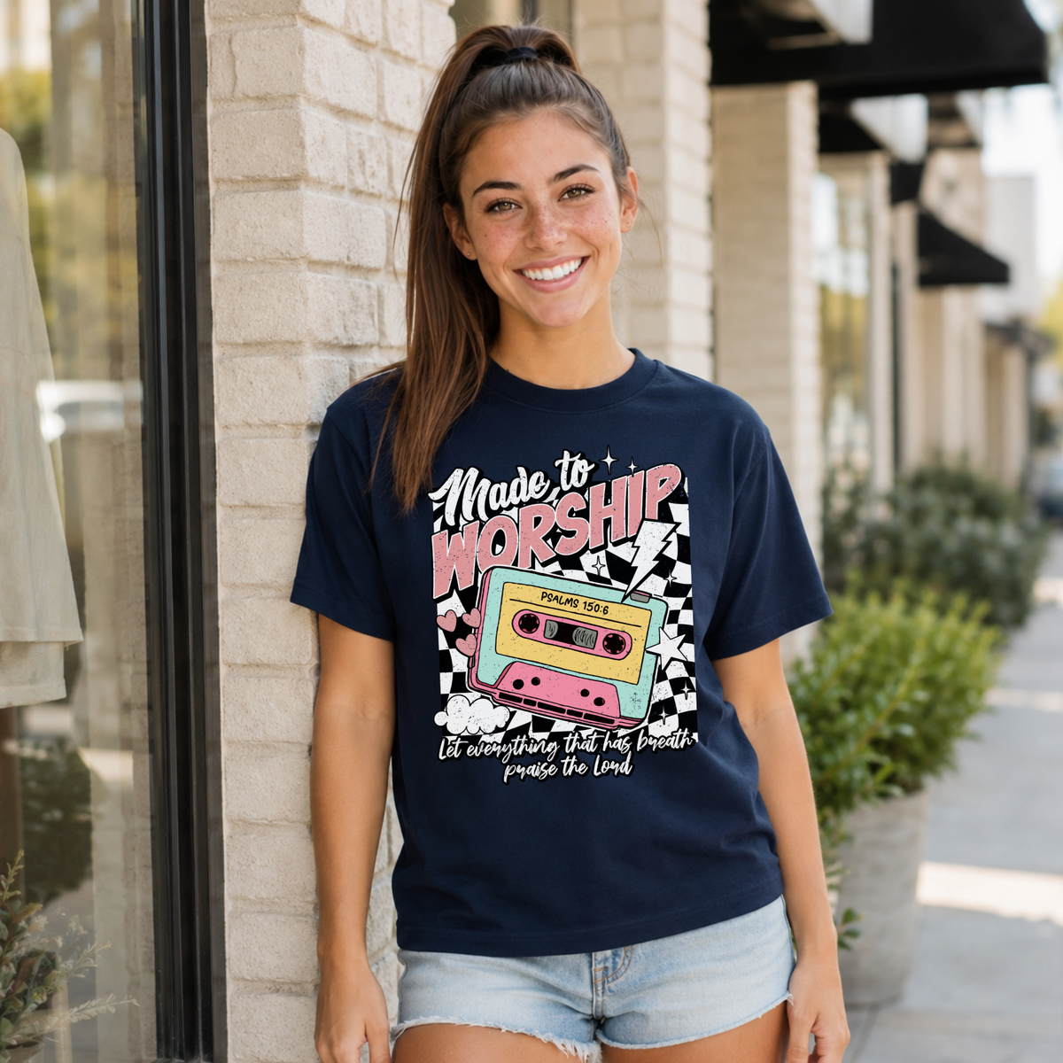 Woman wearing a navy Made to Worship T-shirt with a retro cassette graphic outside a storefront.