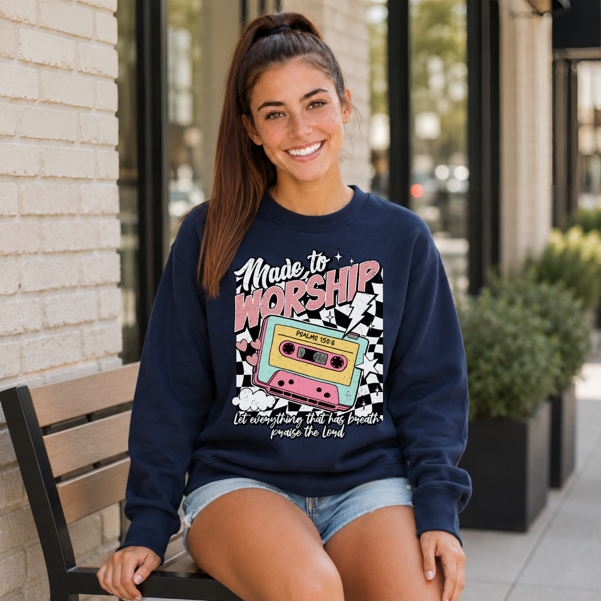 Woman wearing a navy Made to Worship crewneck sweatshirt with a retro cassette graphic while seated outside a storefront.