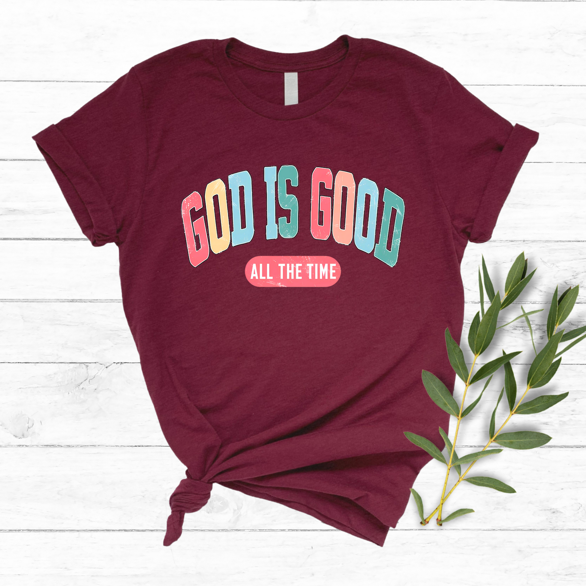 Flat lay of a Maroon T-shirt with 'God Is Good All the Time' in colorful text, perfect for Easter and Christian apparel.