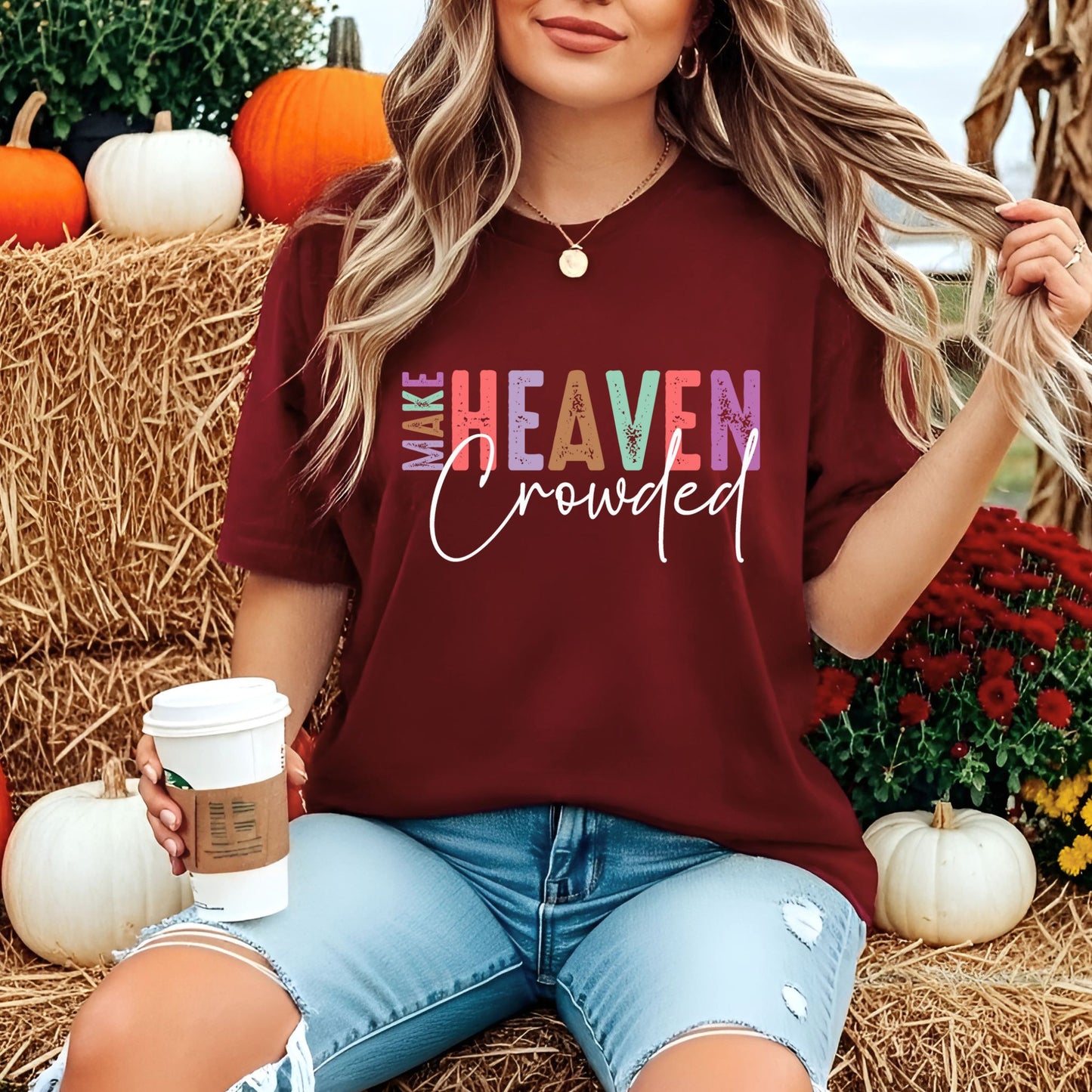 Woman in maroon Make Heaven Crowded graphic T-shirt sitting near hay bales and pumpkins