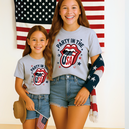 Two girls wearing matching “Party in the USA” shirts with patriotic lips graphic and denim shorts. Standing in front of an American flag, perfect for sisters, cousins, or friends celebrating the 4th of July or Memorial Day.

