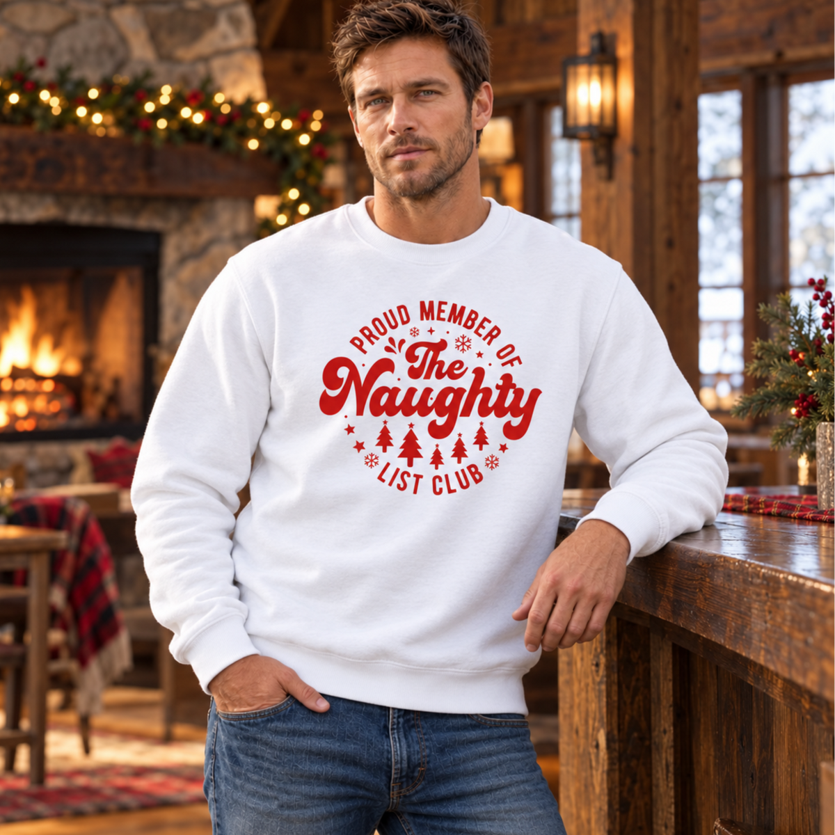 Man wearing a white crewneck sweatshirt with a red Proud Member of the Naughty List Club design beside a cabin fireplace.