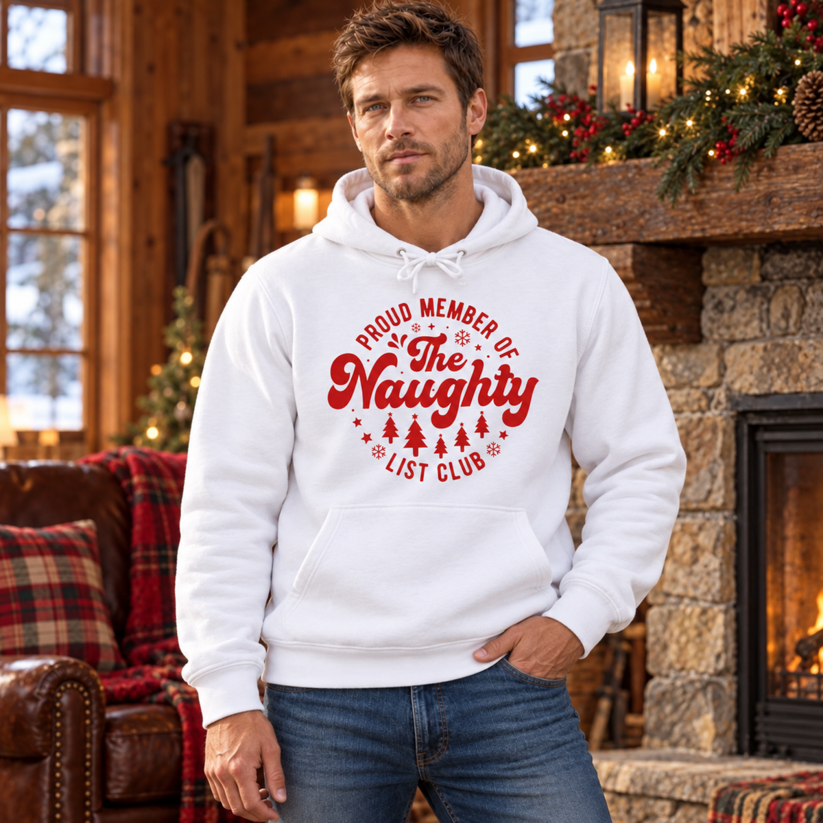 Man wearing a white hoodie with a red Proud Member of the Naughty List Club design in a rustic cabin with a fireplace.
