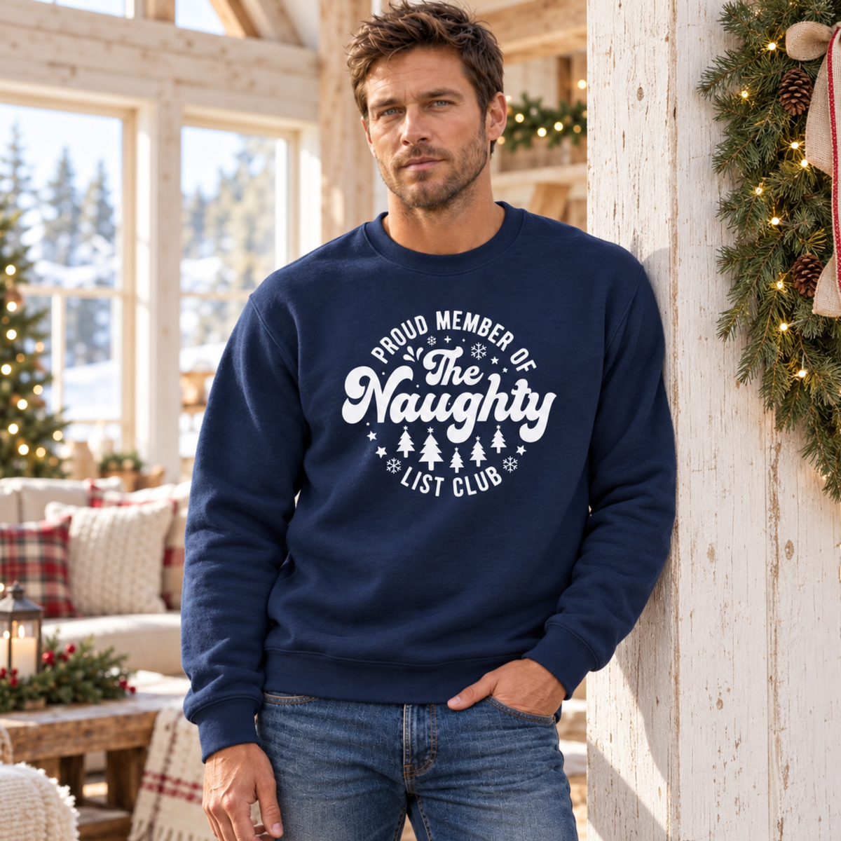 Man wearing a navy crewneck sweatshirt with a white Proud Member of the Naughty List Club design in a festive living room.