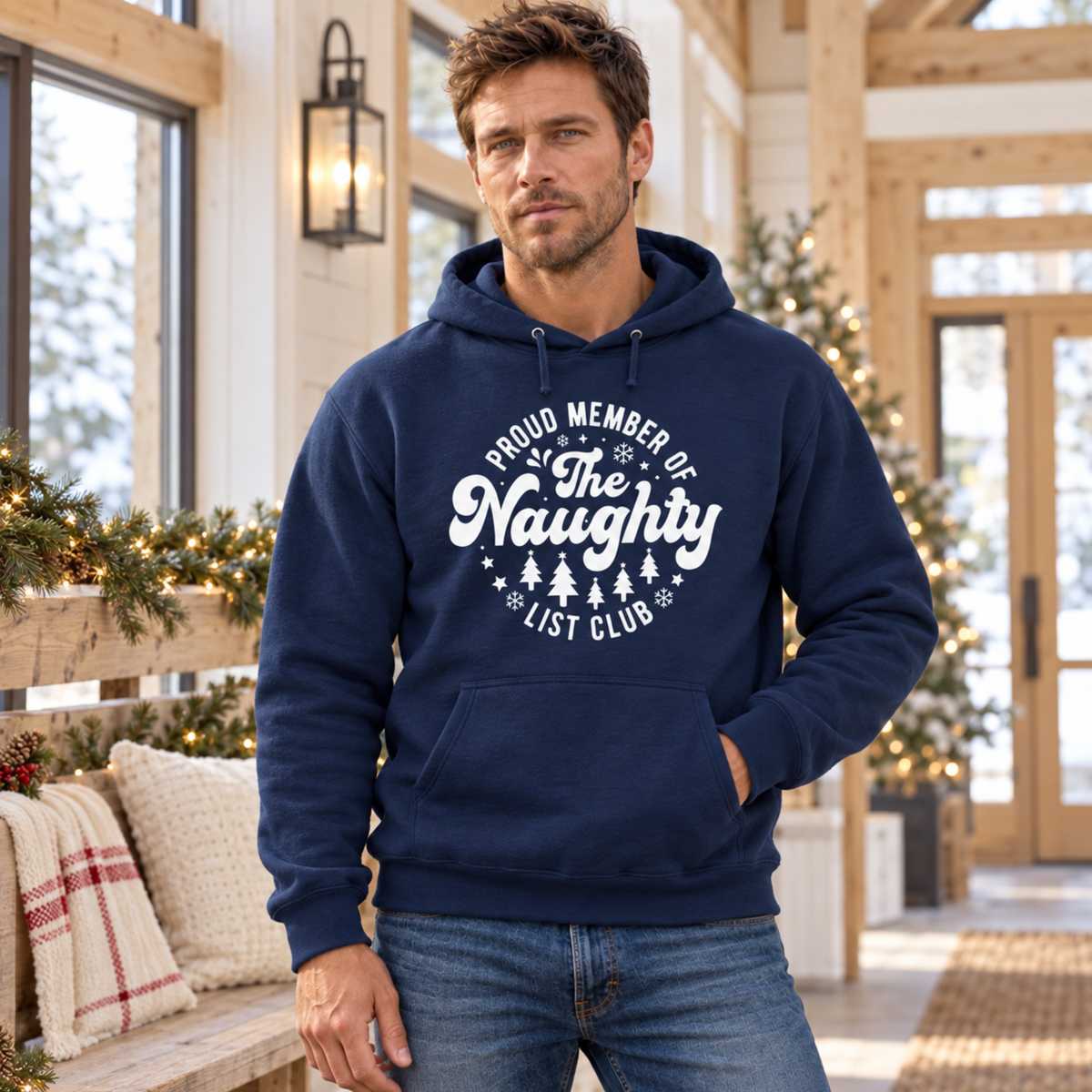 Man wearing a navy hoodie with a white Proud Member of the Naughty List Club design in a decorated holiday cabin.