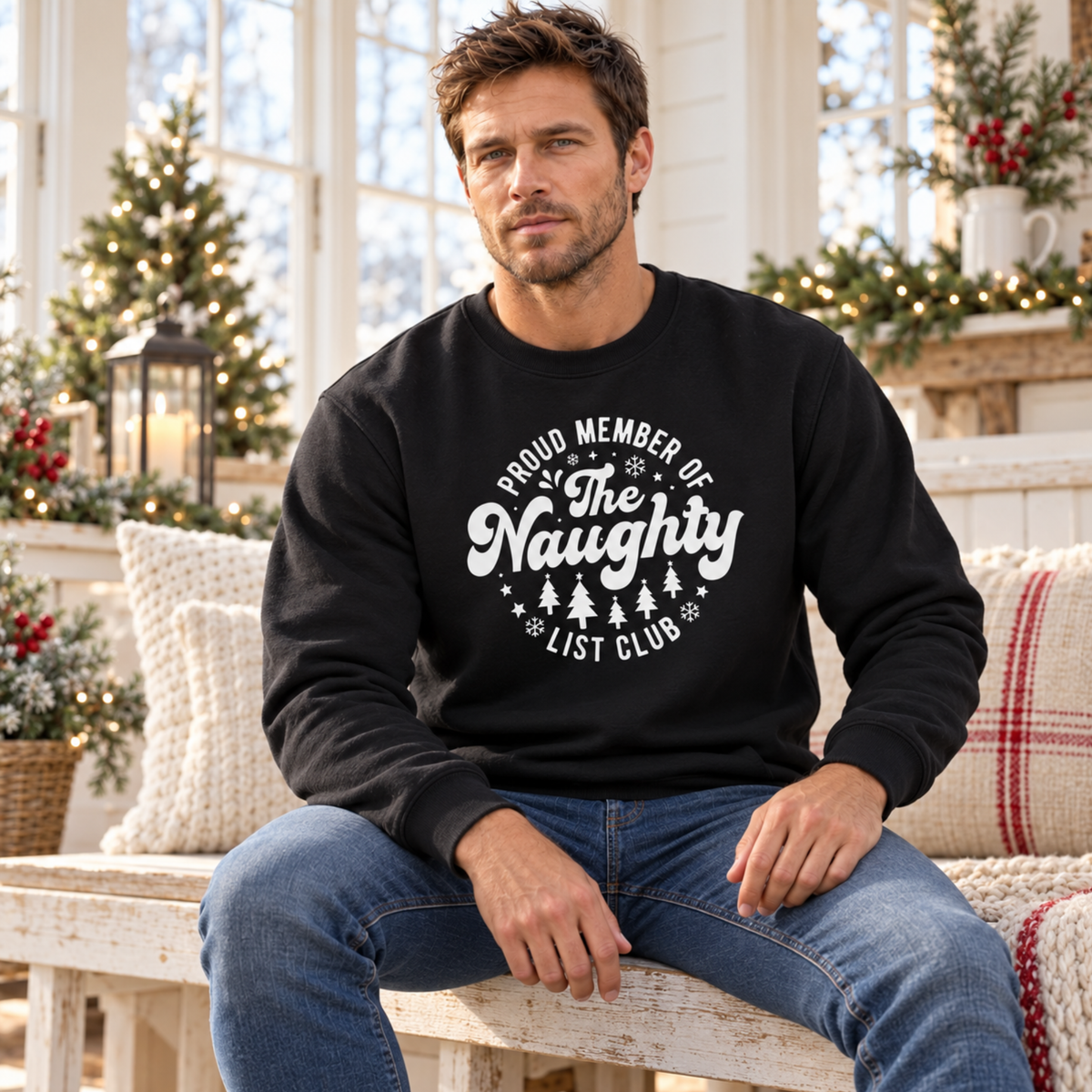 Man wearing a black crewneck sweatshirt with a white Proud Member of the Naughty List Club design while seated in a Christmas room.