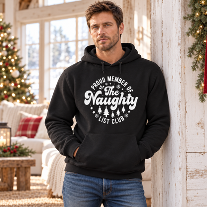 Man wearing a black hoodie with a white Proud Member of the Naughty List Club design beside a Christmas tree.