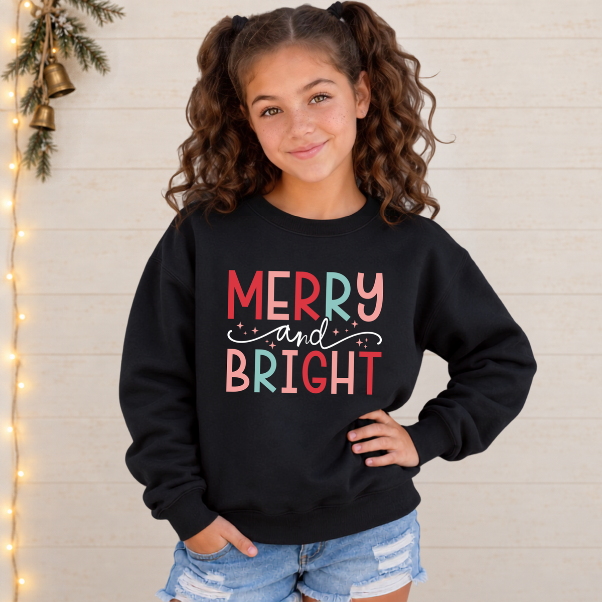 Girl wearing a black youth sweatshirt with colorful Merry and Bright Christmas design against a white wood wall with holiday lights