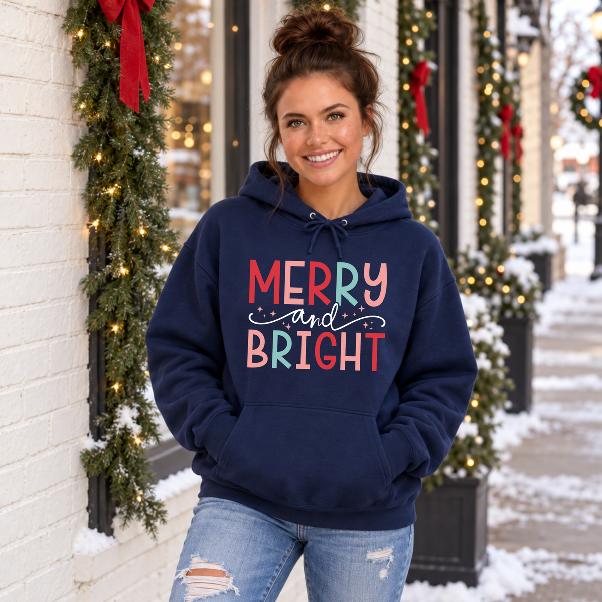 Woman wearing a navy hoodie with colorful Merry and Bright Christmas design outside a snowy decorated storefront