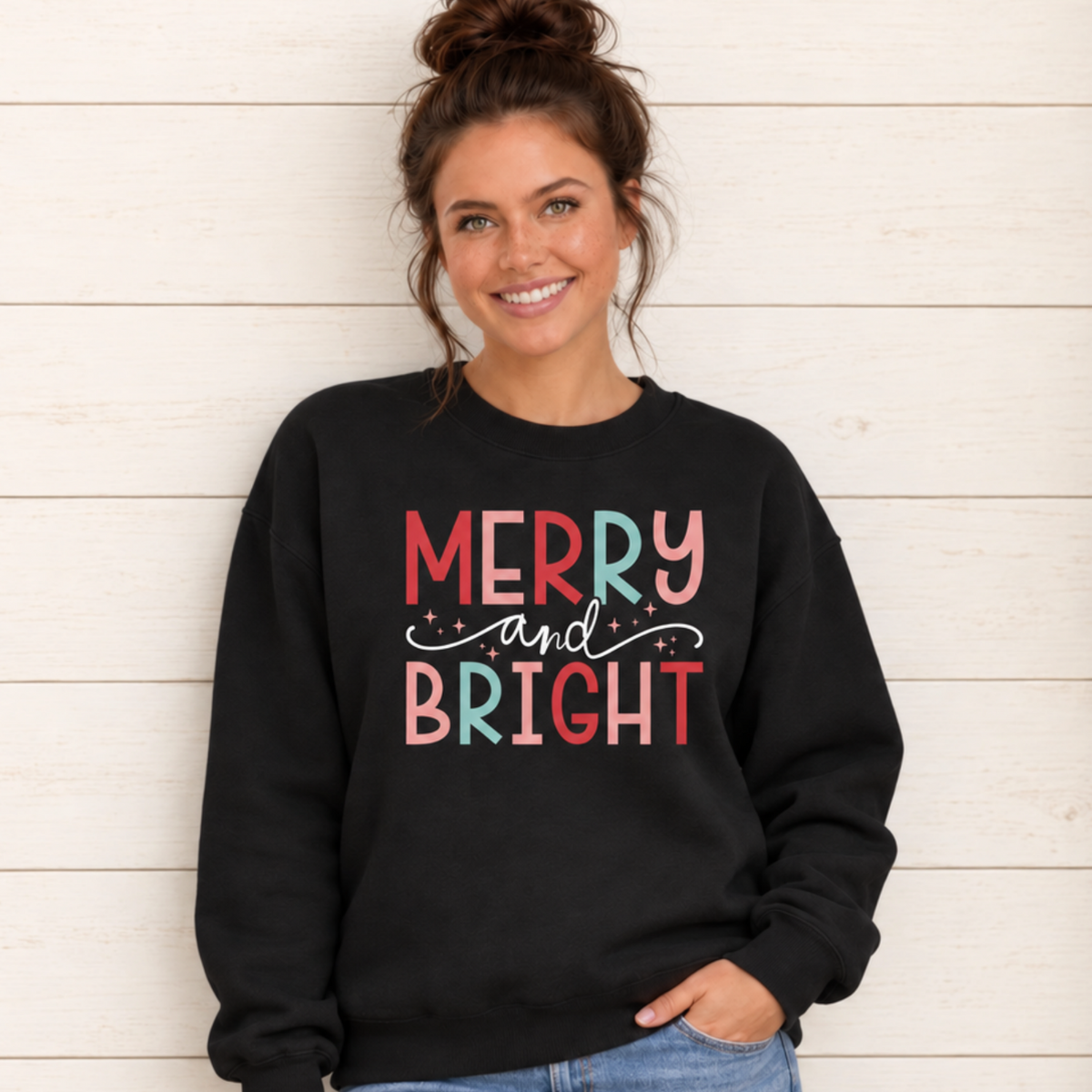 Woman wearing a black sweatshirt with colorful Merry and Bright Christmas design against a white wood wall with holiday greenery