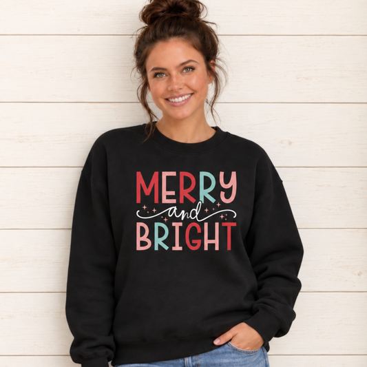 Woman wearing a black sweatshirt with colorful Merry and Bright Christmas design against a white wood wall with holiday greenery