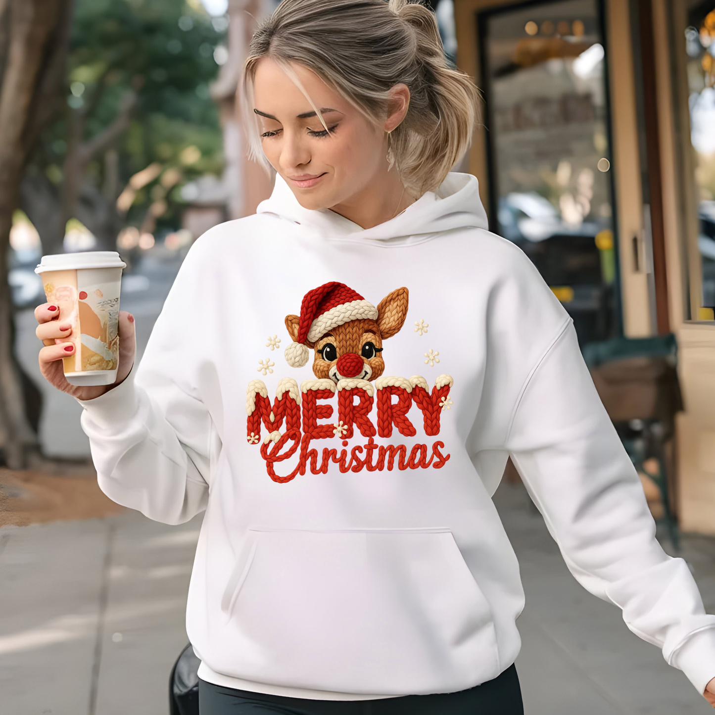 White hoodie with faux crochet Merry Christmas reindeer design worn by woman holding a coffee cup outdoors
