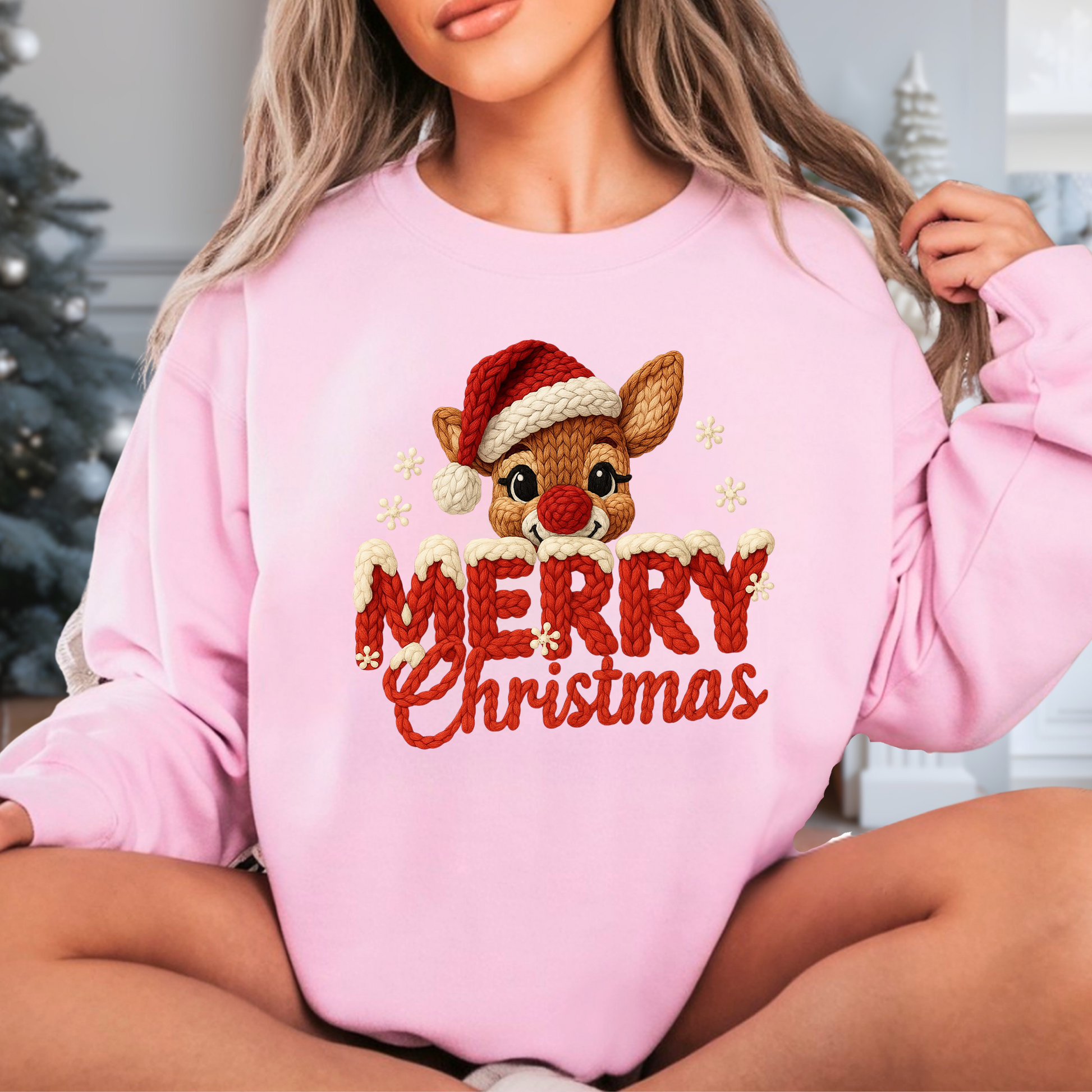 Woman wearing light pink Merry Christmas reindeer chunky knit look sweatshirt sitting by Christmas tree