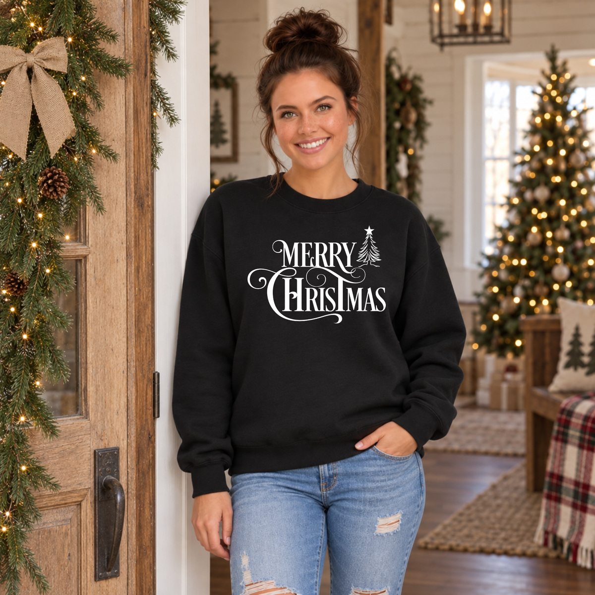 Model in a black Merry Christmas sweatshirt beside a holiday doorway and lit Christmas tree.