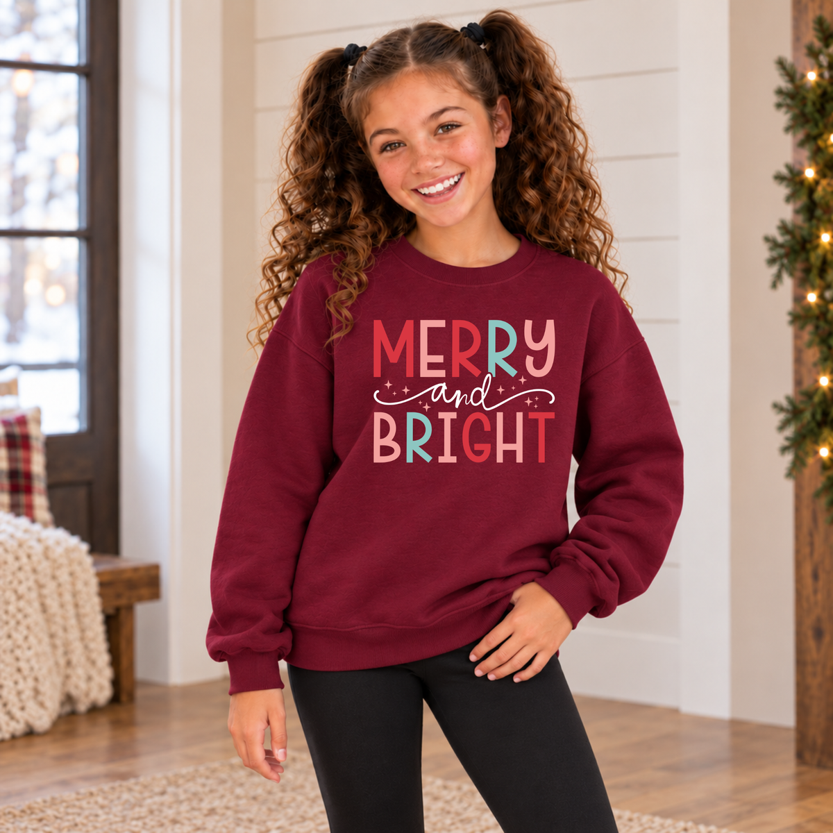 Girl wearing a maroon youth sweatshirt with colorful Merry and Bright Christmas design in a cozy holiday room