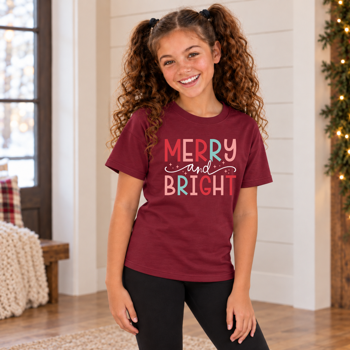 Girl wearing a maroon youth T-shirt with colorful Merry and Bright Christmas design in a cozy holiday room