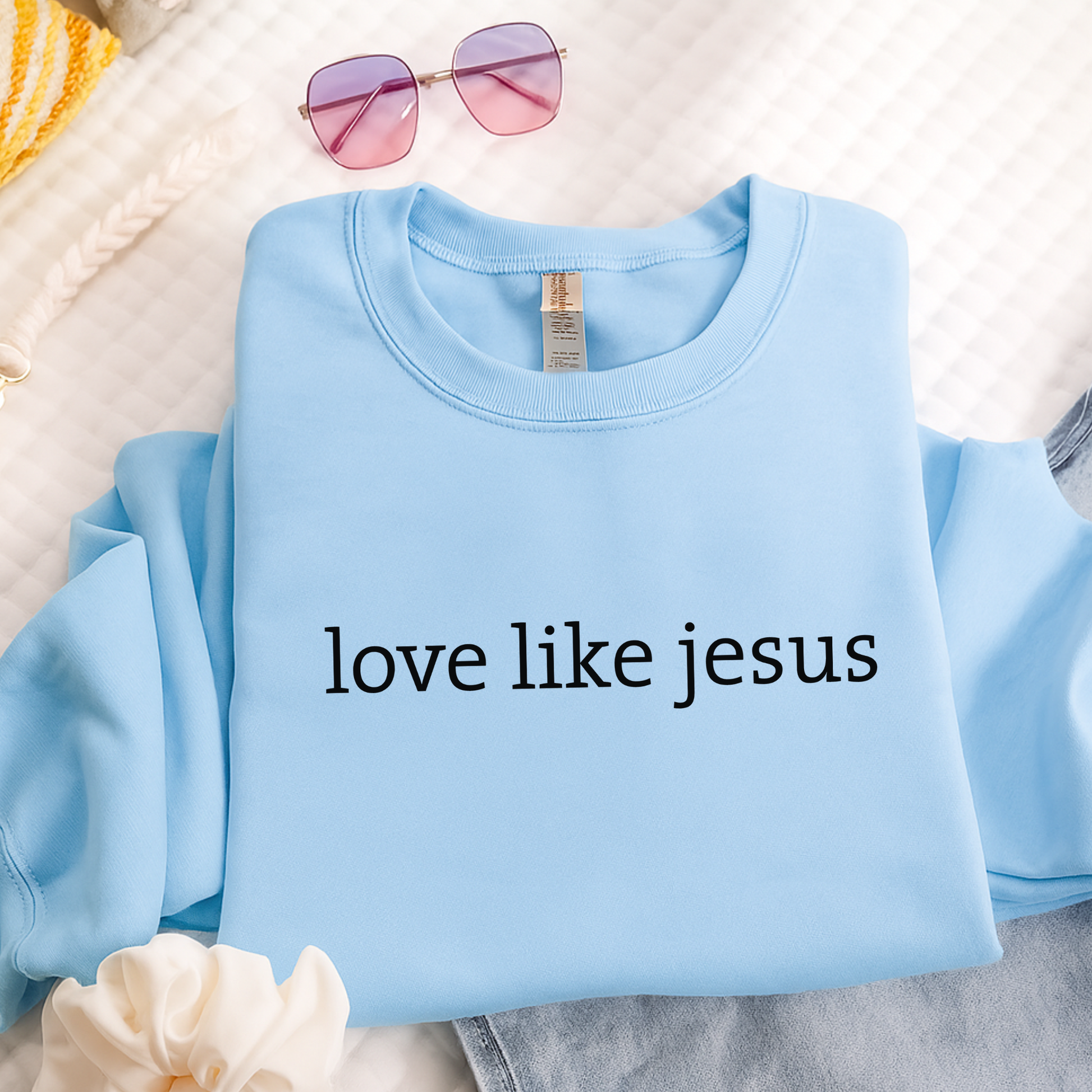 Folded blue “Love Like Jesus” sweatshirt on bed with sunglasses