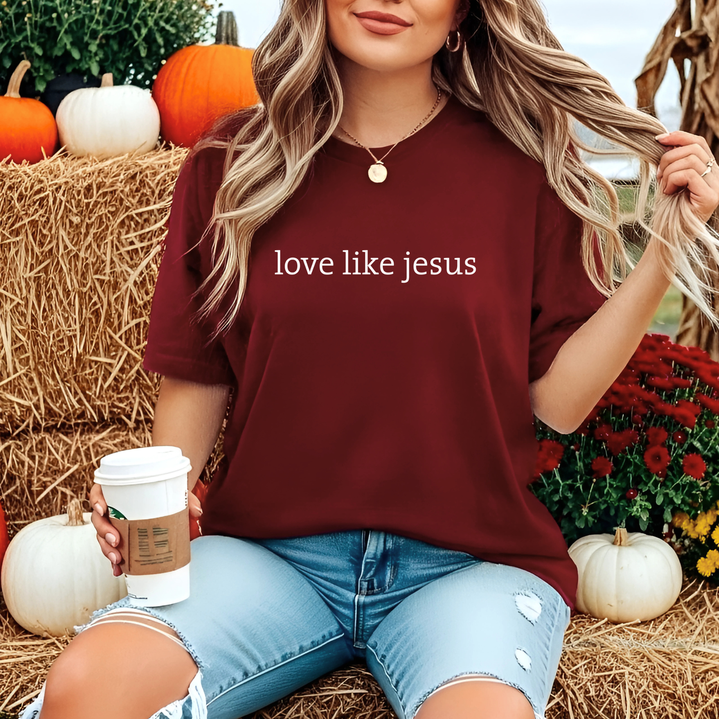 Woman wearing maroon “Love Like Jesus” T-shirt sitting in pumpkin patch with latte