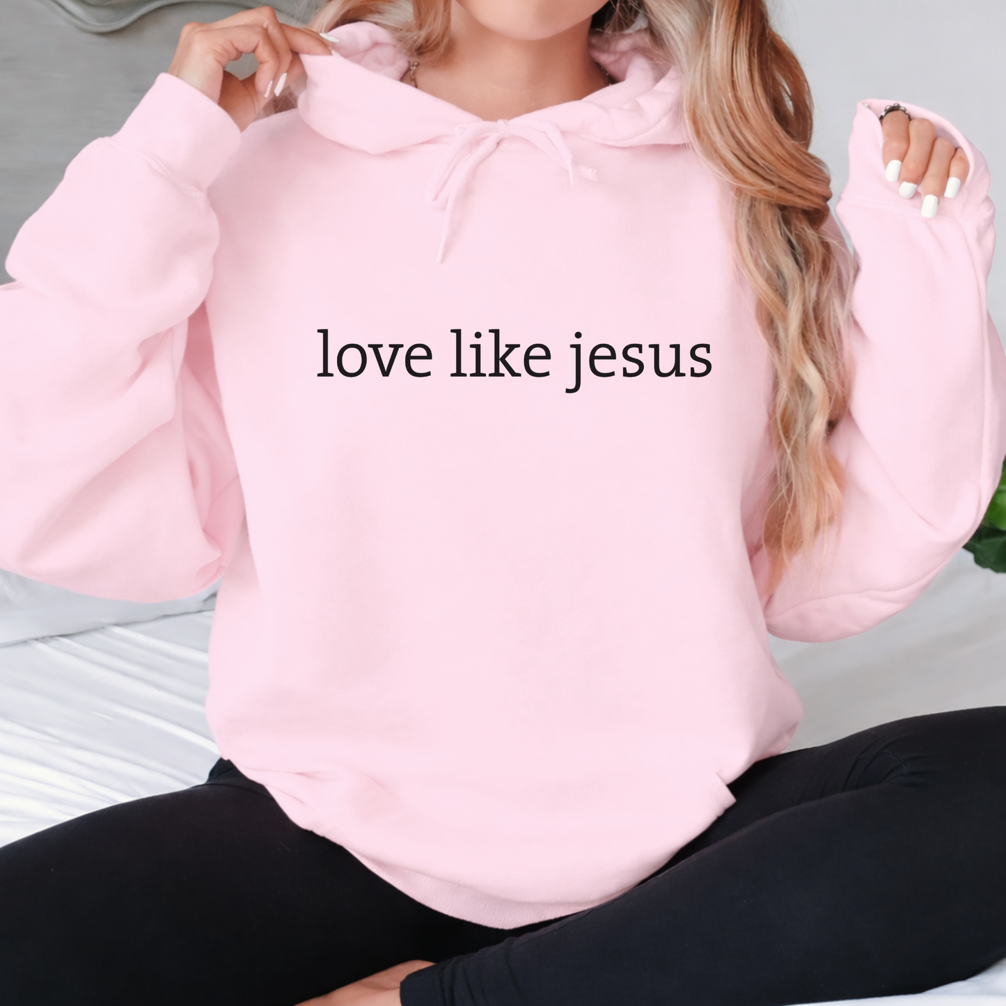 Lifestyle image of woman wearing light pink hoodie with “love like jesus” text while seated