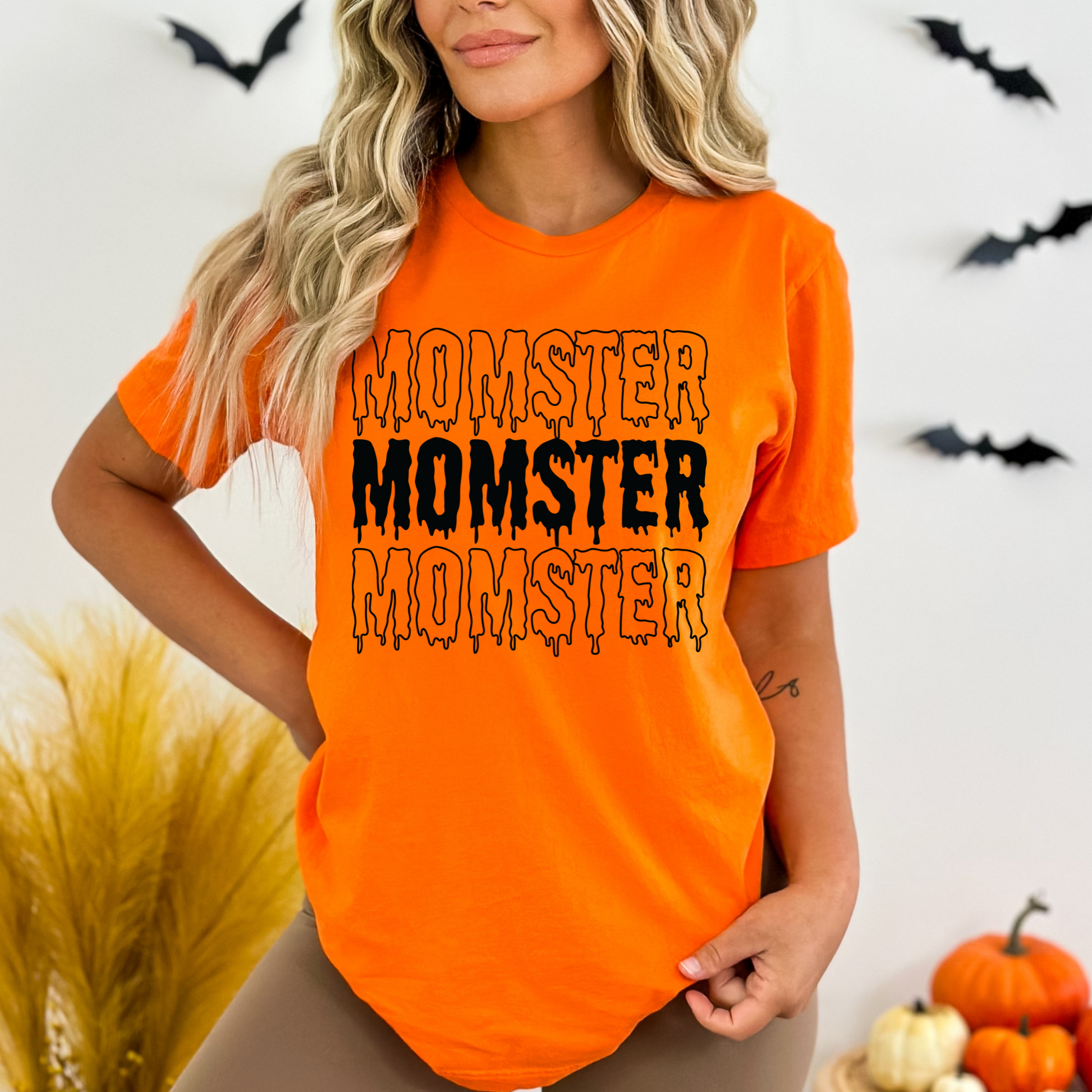 Woman wearing an orange t-shirt with 'MOMSTER' text, Halloween-themed background with pumpkins and bats.