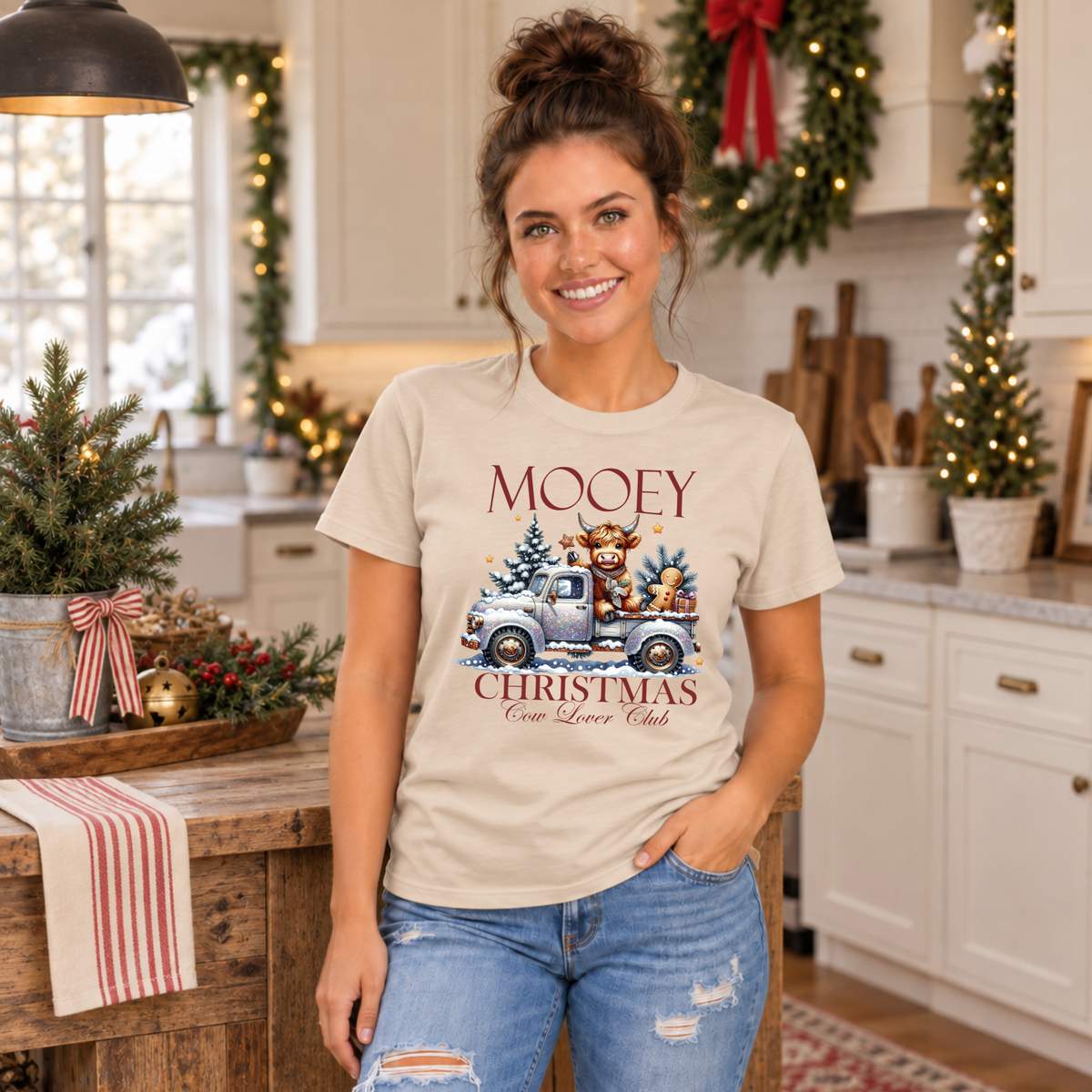 Woman wearing a sand T-shirt with Mooey Christmas Highland cow and truck graphic in a festive kitchen.