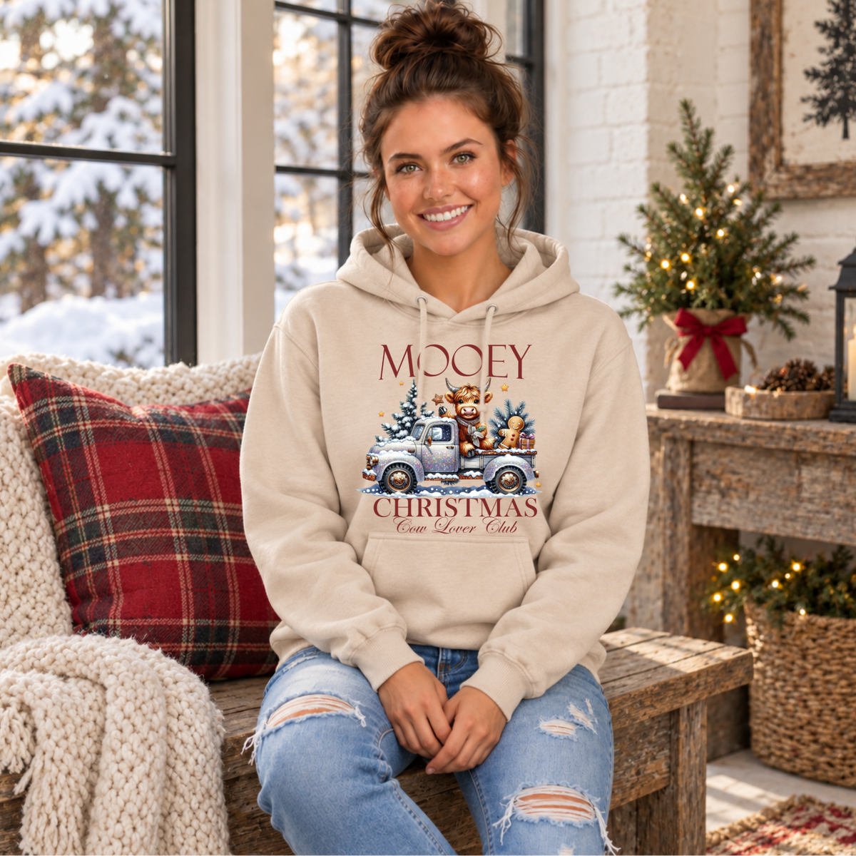 Woman seated in a sand hoodie with Mooey Christmas Highland cow and vintage truck graphic.