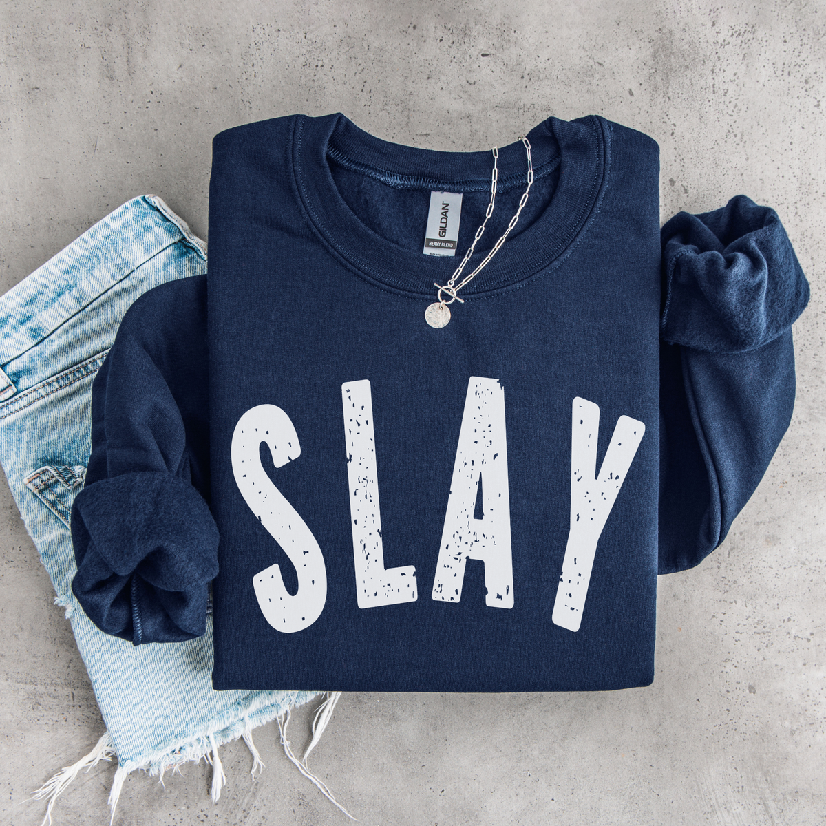 Navy SLAY crewneck sweatshirt flat lay with bold white distressed lettering, styled with denim, necklace, and matching scrunchies.