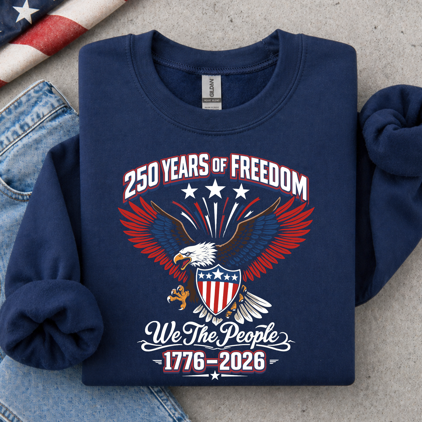 Navy blue America 250 sweatshirt with eagle design and text on a gray surface with denim jeans and American flag.