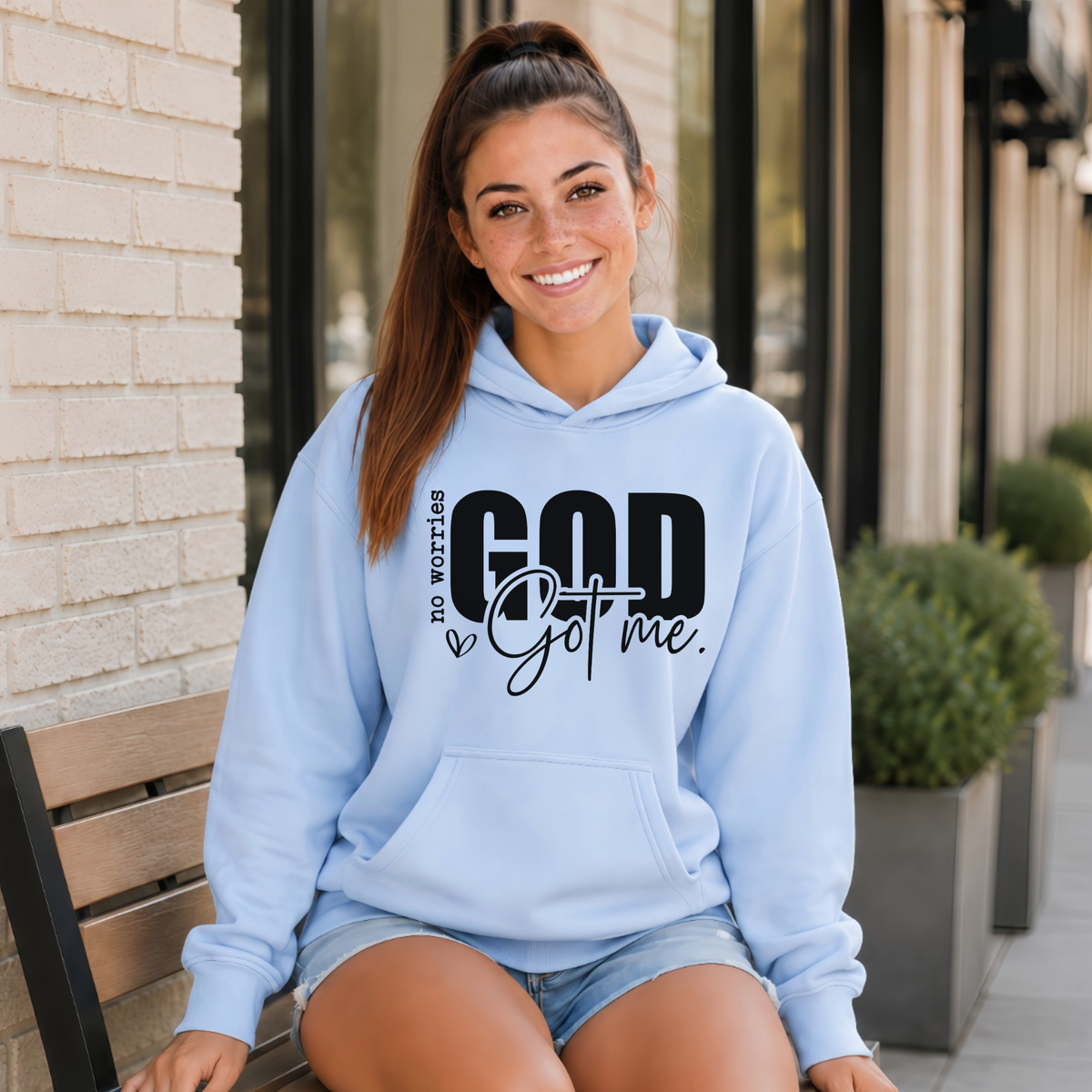 Woman wearing a light blue No Worries God Got Me hoodie with bold black graphic lettering outside a storefront.