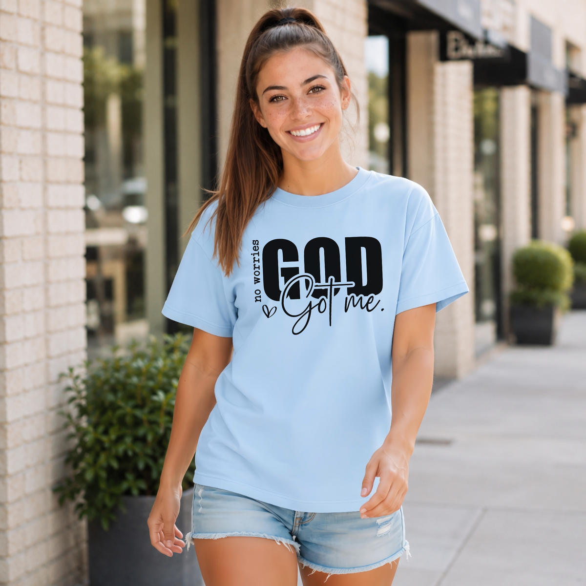 Woman wearing a light blue No Worries God Got Me T-shirt with bold black graphic lettering outside a storefront.