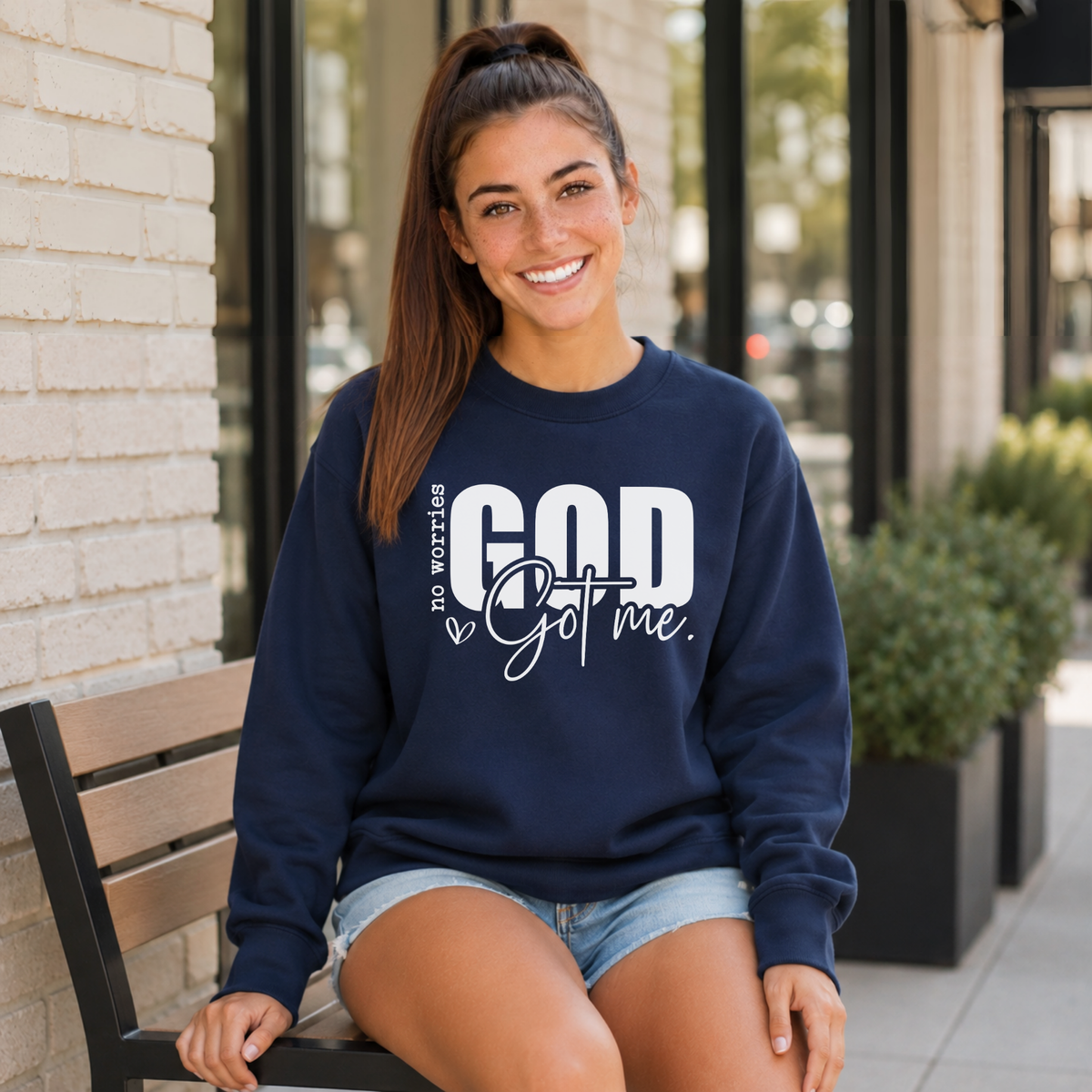 Woman wearing a navy No Worries God Got Me crewneck sweatshirt with bold white graphic lettering while seated outside.