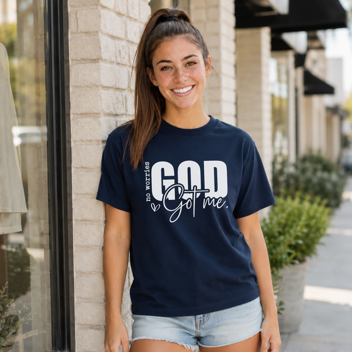 Woman wearing a navy No Worries God Got Me T-shirt with bold white graphic lettering outside a storefront.