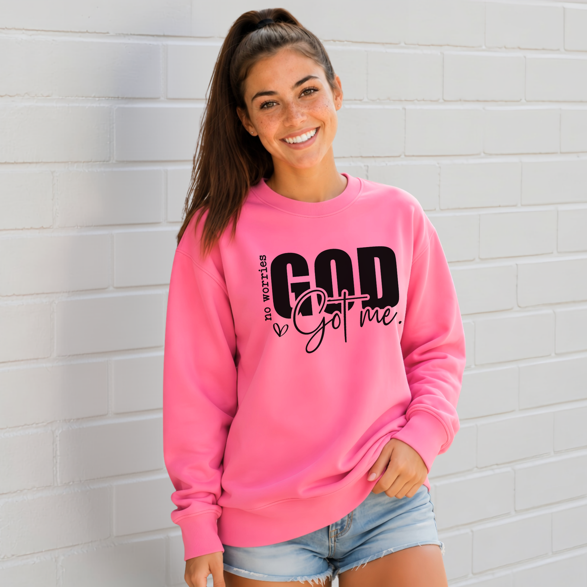 Woman wearing a Bright Pink No Worries God Got Me crewneck sweatshirt with bold black graphic lettering against a white brick wall.