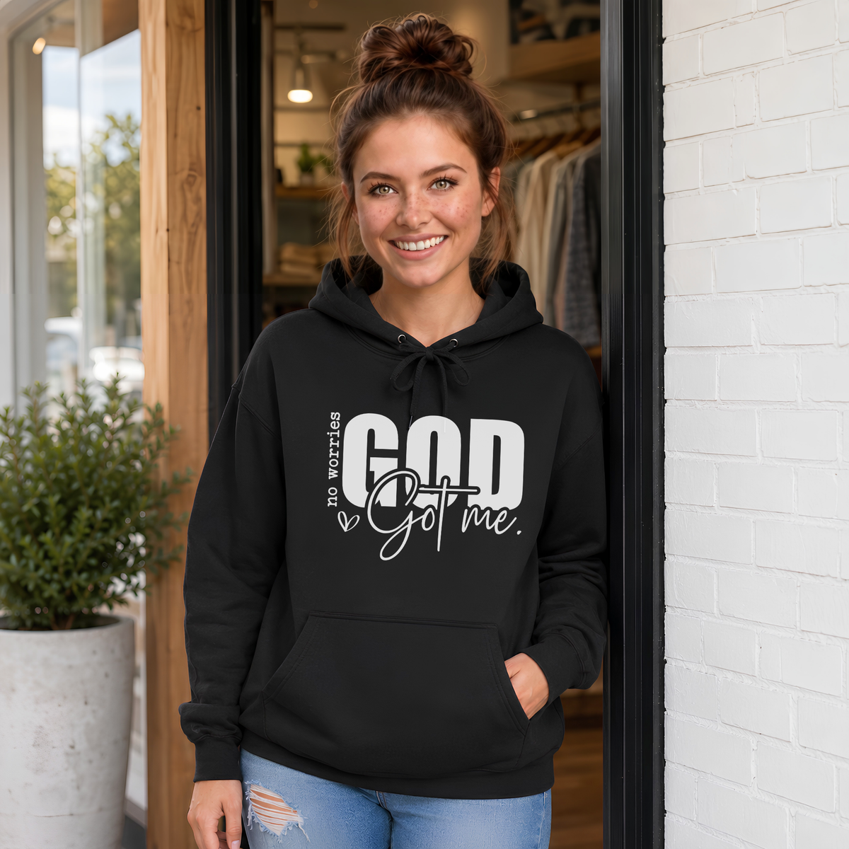 Woman wearing a black No Worries God Got Me hoodie with bold white graphic lettering outside a storefront.