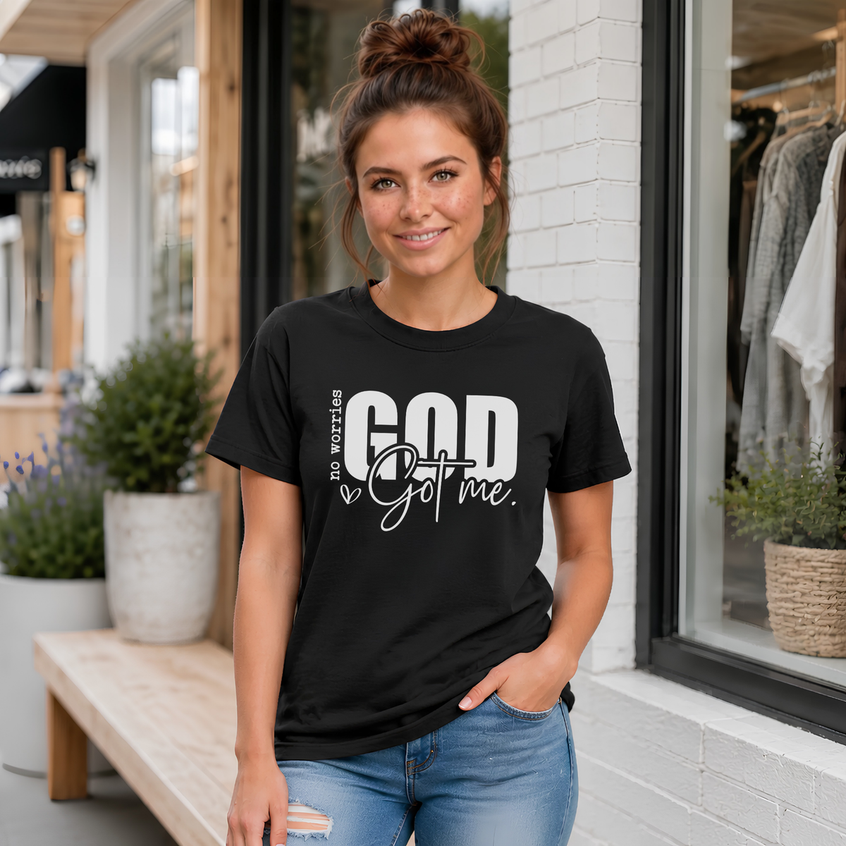Woman wearing a black No Worries God Got Me T-shirt with bold white graphic lettering outside a boutique.
