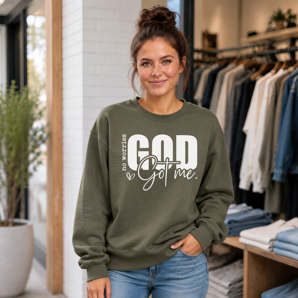 Woman wearing a military green No Worries God Got Me crewneck sweatshirt with bold white graphic lettering inside a boutique.