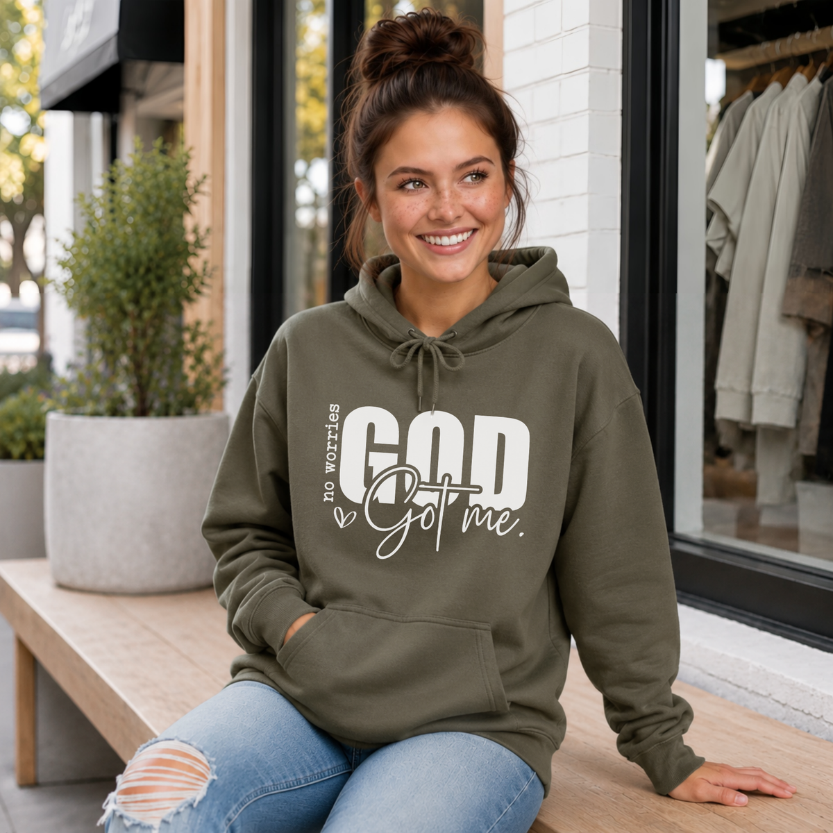 Woman wearing a military green No Worries God Got Me hoodie with bold white graphic lettering outside a boutique.