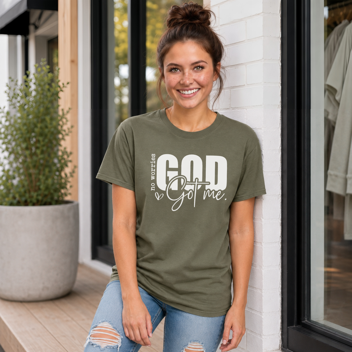 Woman wearing a military green No Worries God Got Me T-shirt with bold white graphic lettering outside a boutique.