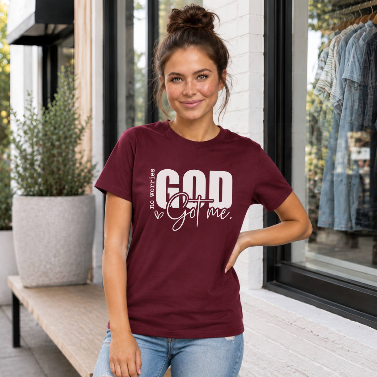 Woman wearing a maroon No Worries God Got Me T-shirt with bold white graphic lettering outside a boutique.