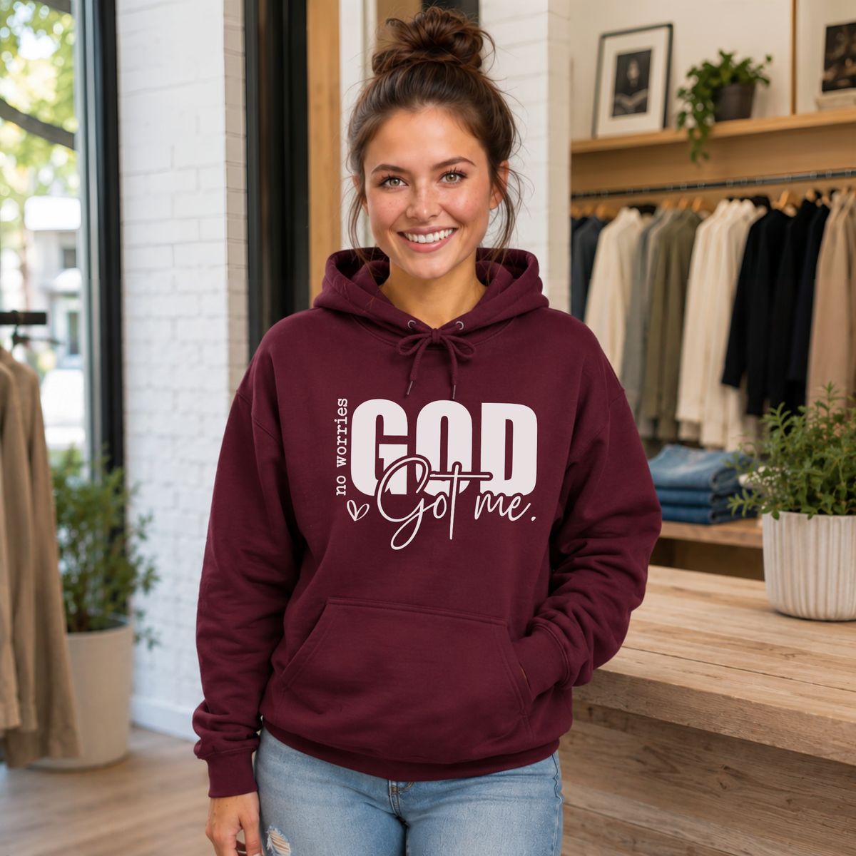 Woman wearing a maroon No Worries God Got Me hoodie with bold white graphic lettering inside a boutique.